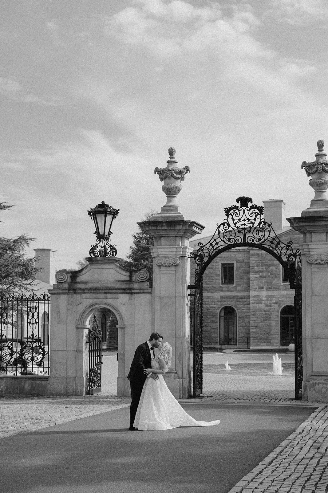 A black and white photograph of a wedding couple standing under an ornate gate outside a historic building with fountains, with the groom leaning in to kiss the bride.