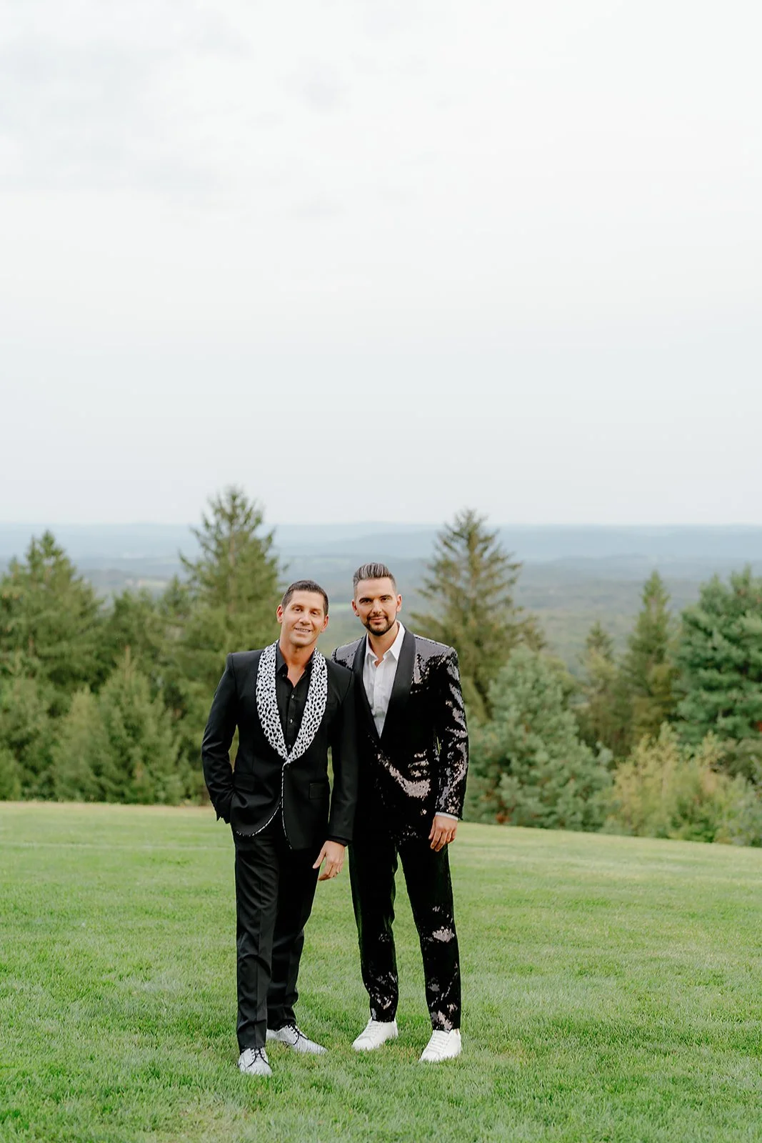Two men in suits standing outdoors on a grassy field with trees in the background, smiling at the camera.