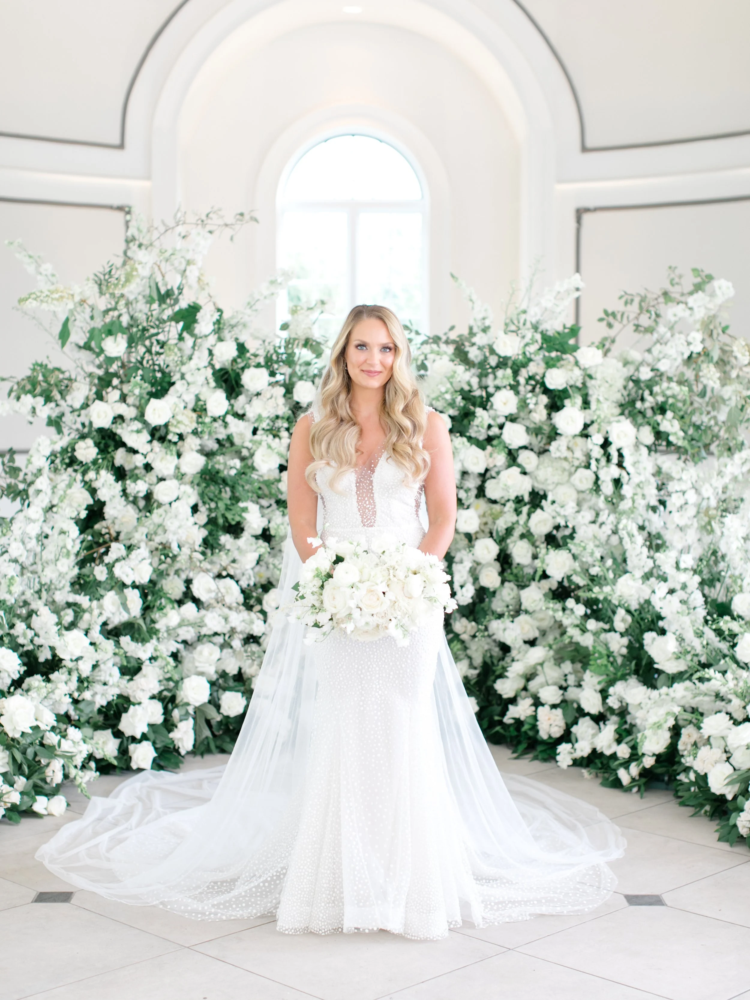 A bride in a white wedding dress holding a bouquet of white flowers, standing in front of an arrangement of white flowers with a large window behind in a bright, elegant room.