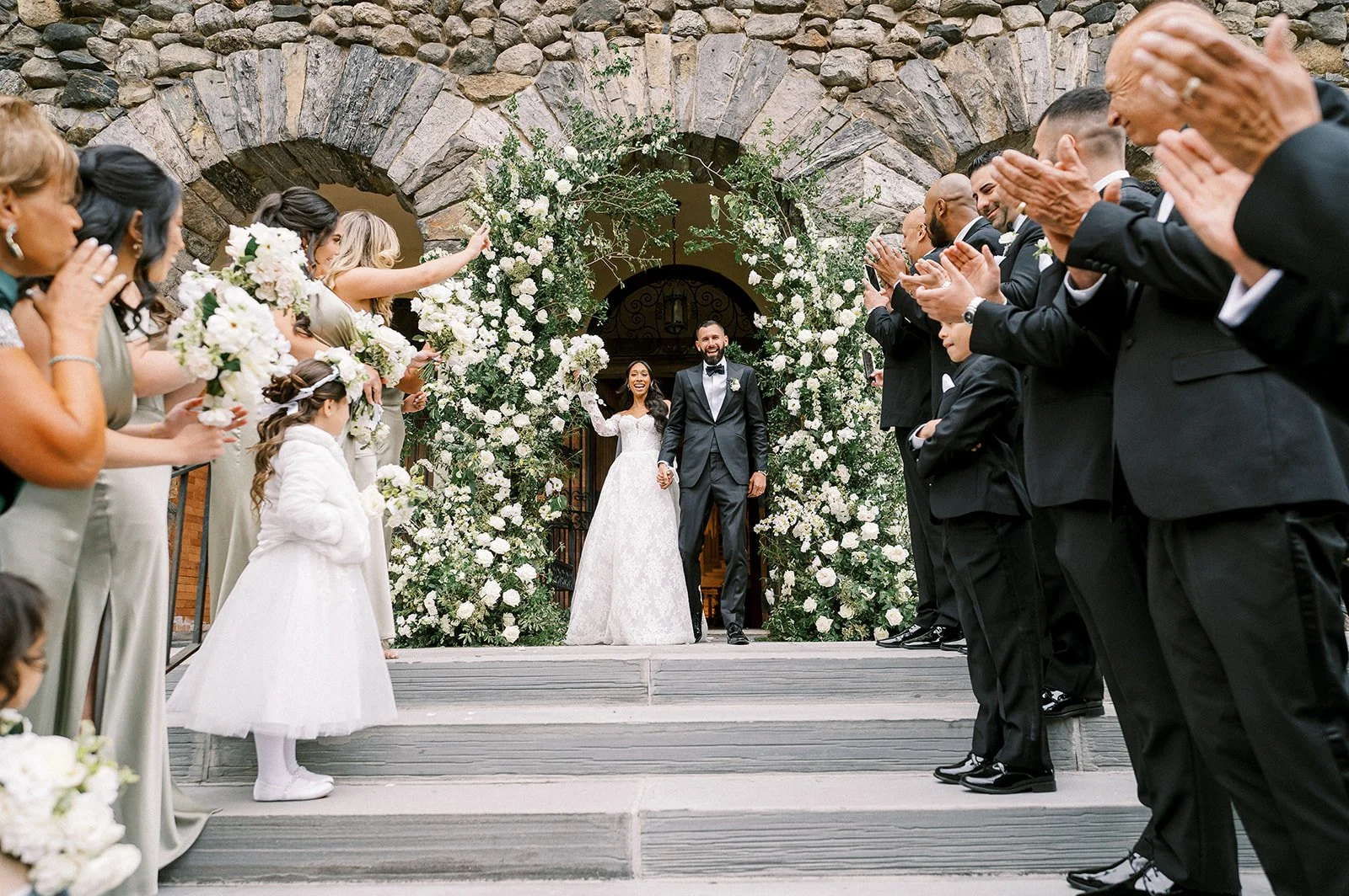 A newlywed couple walks out of a stone building under a floral arch, surrounded by wedding guests. The bride is in a white wedding gown, and the groom in a black tuxedo, holding hands and smiling. The guests are clapping and celebrating the couple's 