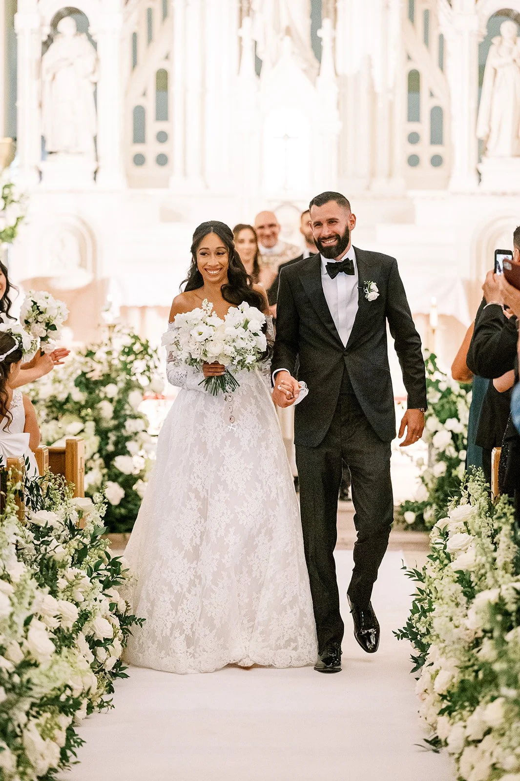 A bride and groom walking down the aisle during a wedding ceremony, surrounded by white flowers and guests in a decorated church or chapel.