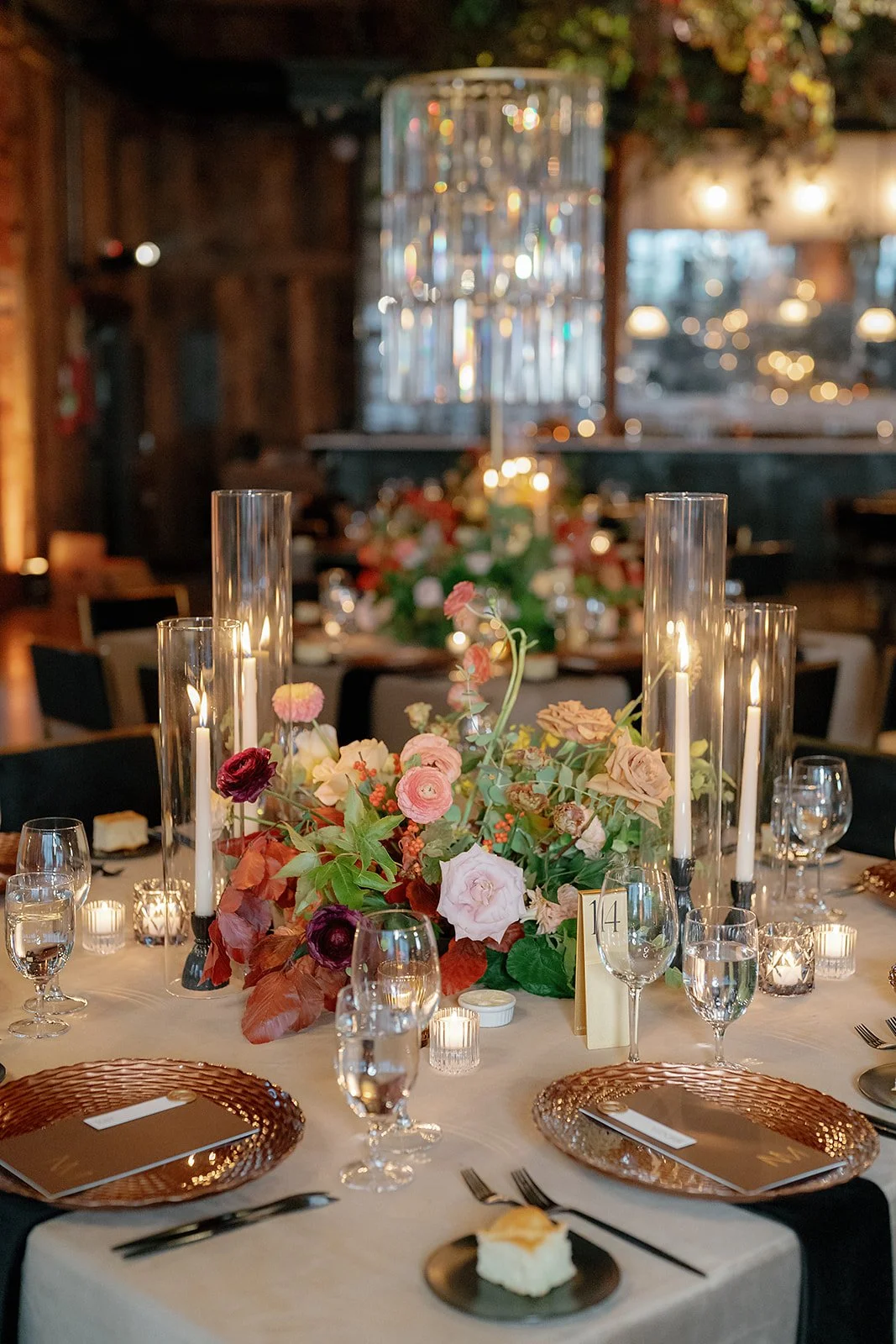 Elegant round table set for a formal event with a flower centerpiece, candles, and place settings, in a warmly lit rustic venue.