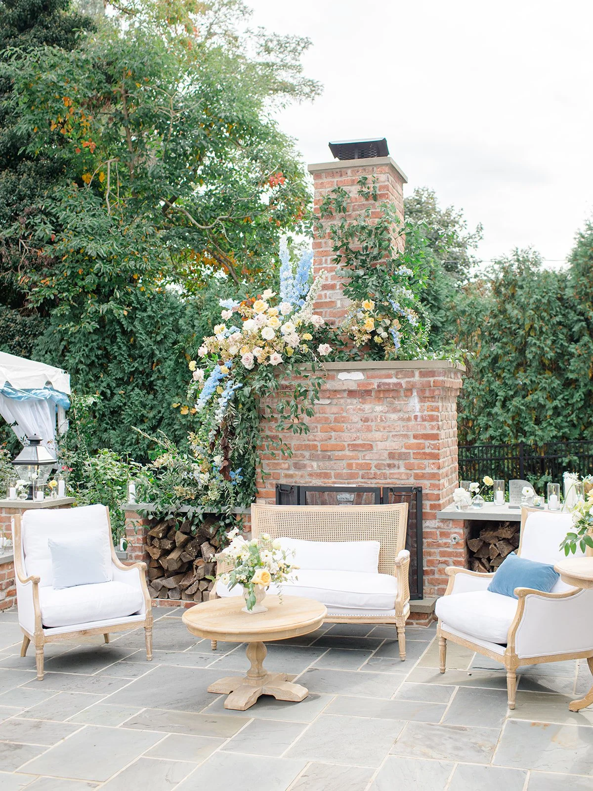 Outdoor patio with white upholstered chairs, a round wooden table with a floral arrangement, a brick fireplace adorned with flowers, and a stack of firewood beneath the fireplace, surrounded by trees.