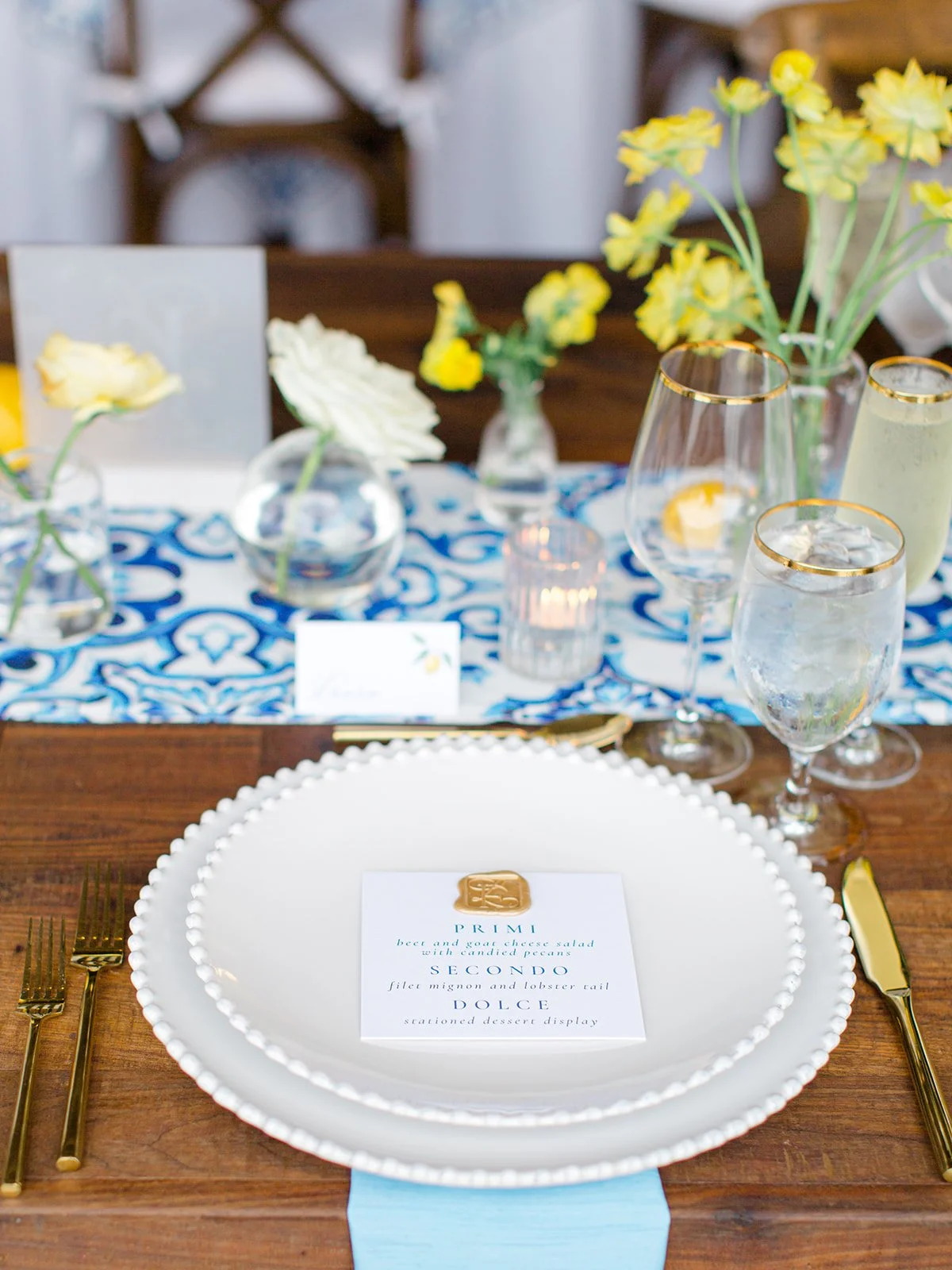 Elegant table setting with a large white beaded-edge plate, gold utensils, a blue table runner, and a menu card with a gold emblem. The background features vases with yellow flowers, water glasses, and a lit candle.