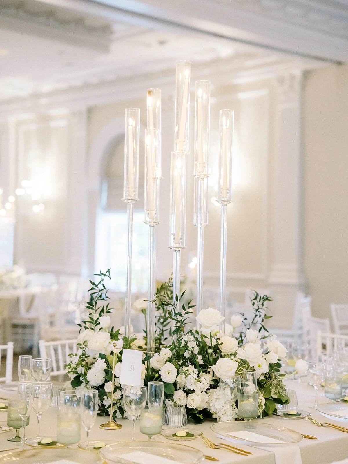 Elegant table centerpiece with tall white candles, surrounded by white flowers and greenery in a formal dining setting.