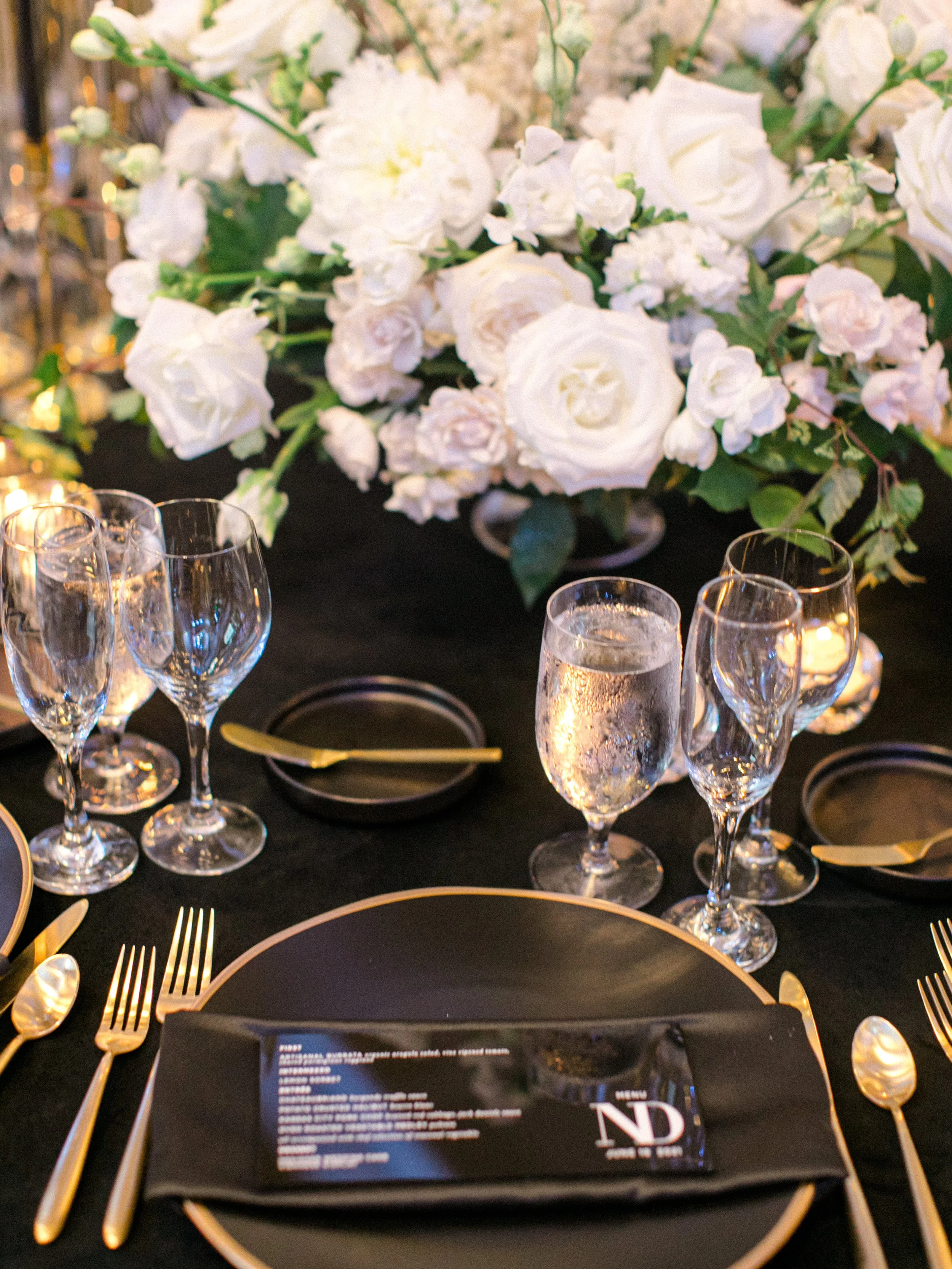 Elegant table settings with black and gold accents, including gold utensils, black plates, and a black napkin with a menu, topped with a large floral centerpiece of white roses and greenery, and glasses of water and champagne on a black tablecloth.