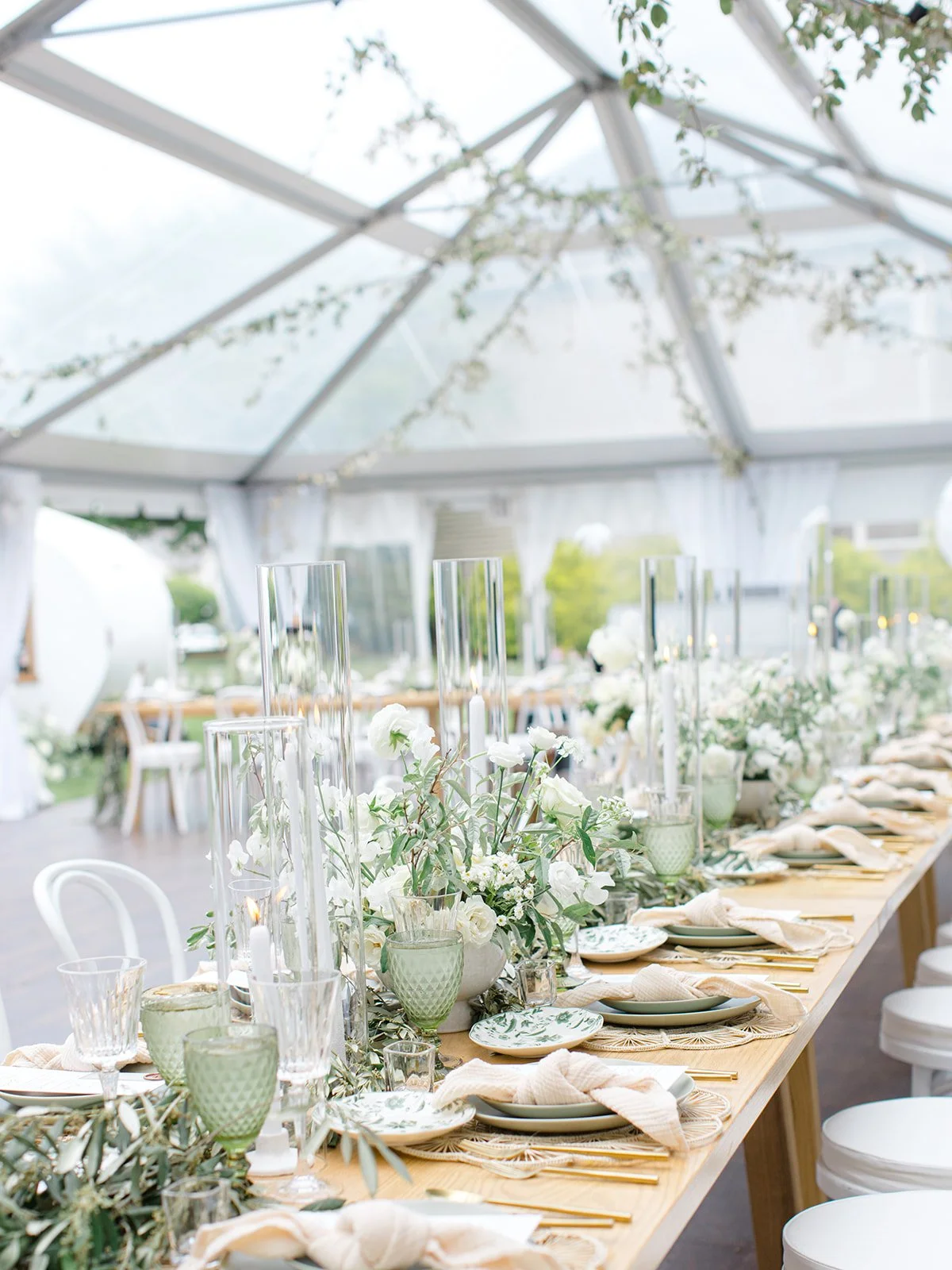 Decorated outdoor dining table with white flowers, green glasses, and candles under a transparent tent with greenery.