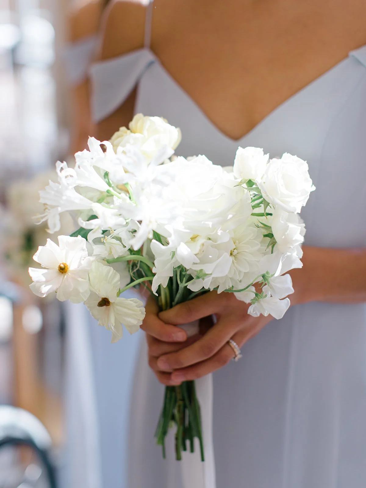 Person holding a bouquet of white flowers, wearing a light-colored dress with thin straps and a ring on their finger.