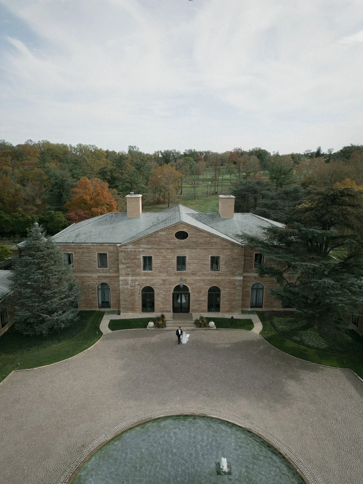 Aerial view of a large stone mansion with a gray metal roof, surrounded by trees and a paved courtyard with a fountain at the front.