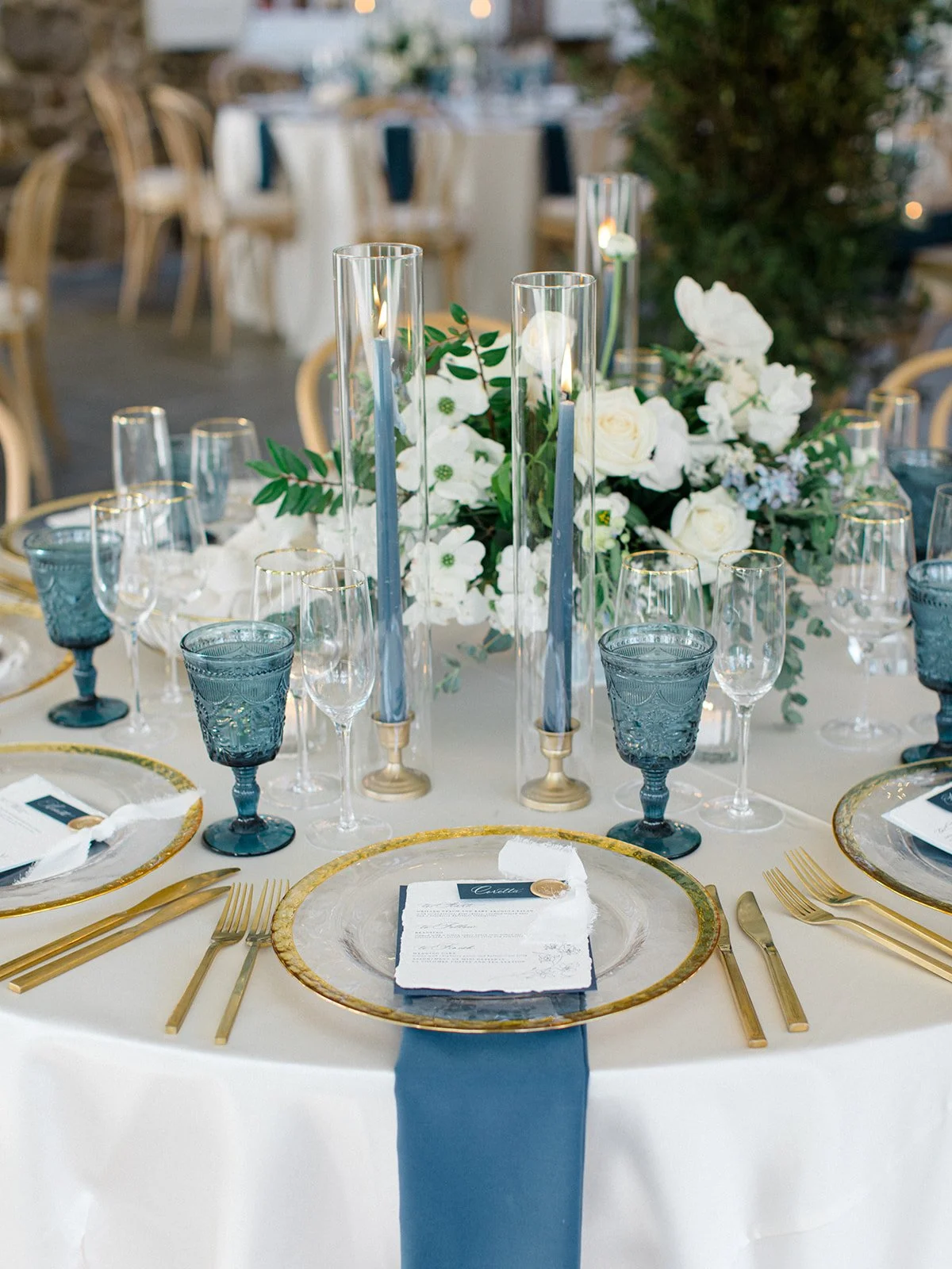 Elegant round table setting with gold-rimmed clear glass plates, gold flatware, blue textured glasses, a white napkin, and a menu card. Centerpiece features tall glass candles and a floral arrangement of white flowers and greenery.