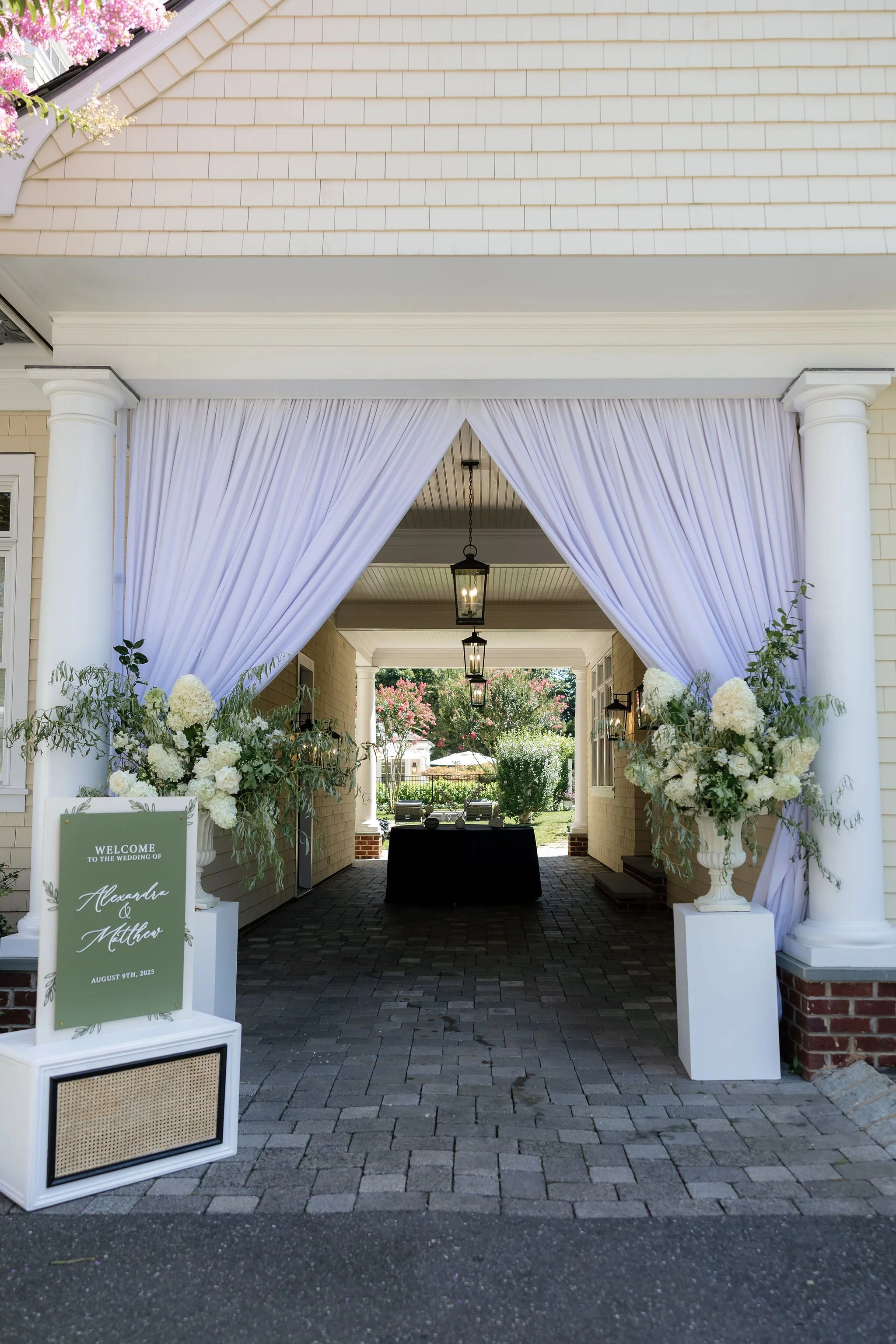 Decorated outdoor wedding entrance with white drapes, floral arrangements, and a welcome sign for Alexandra and Matthew on August 9th, 2023.