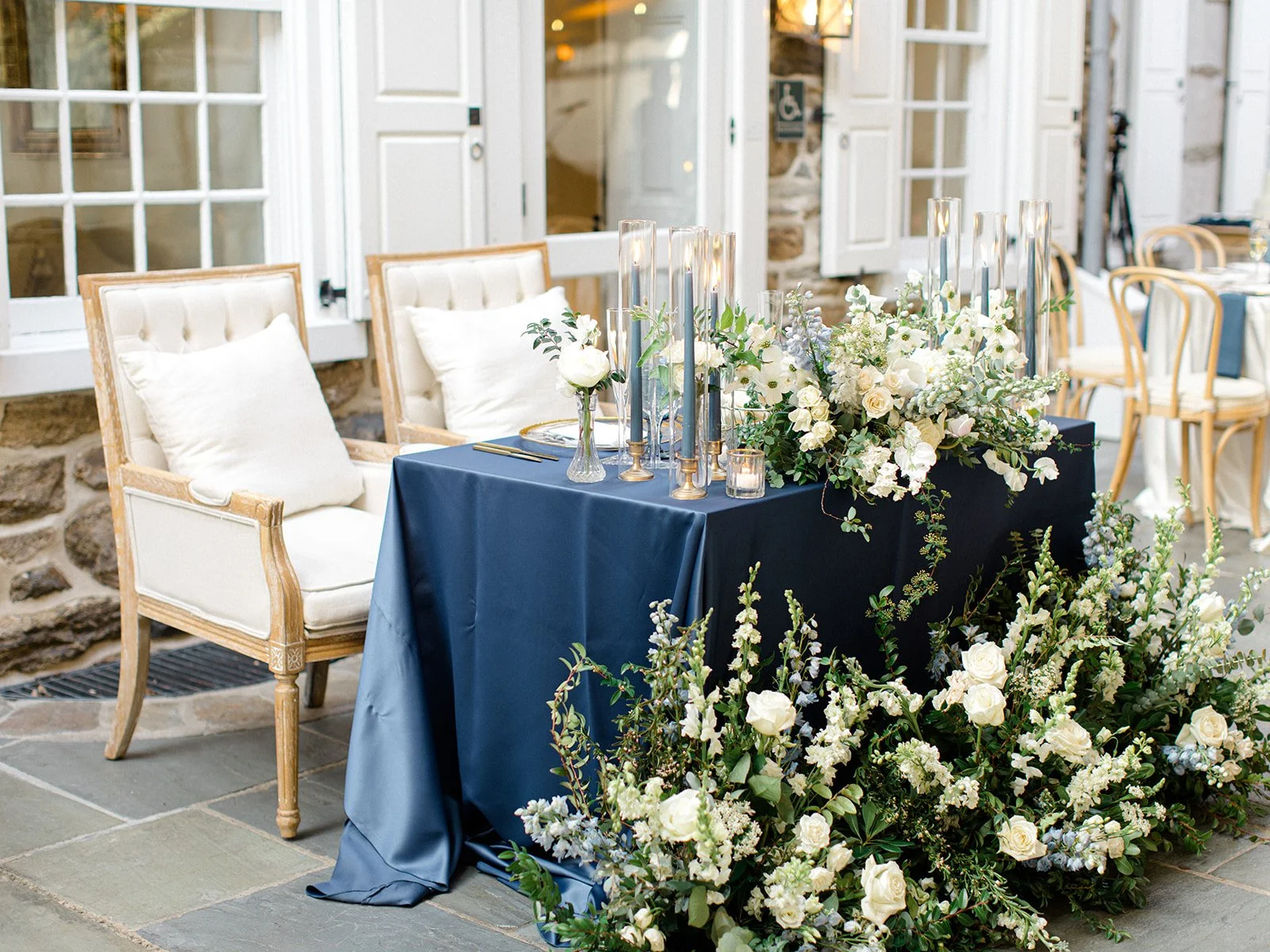 Elegant indoor event with a table draped in navy blue cloth, decorated with white flowers, tall candles, and seating with cream-colored pillows, set against a stone and wood backdrop.