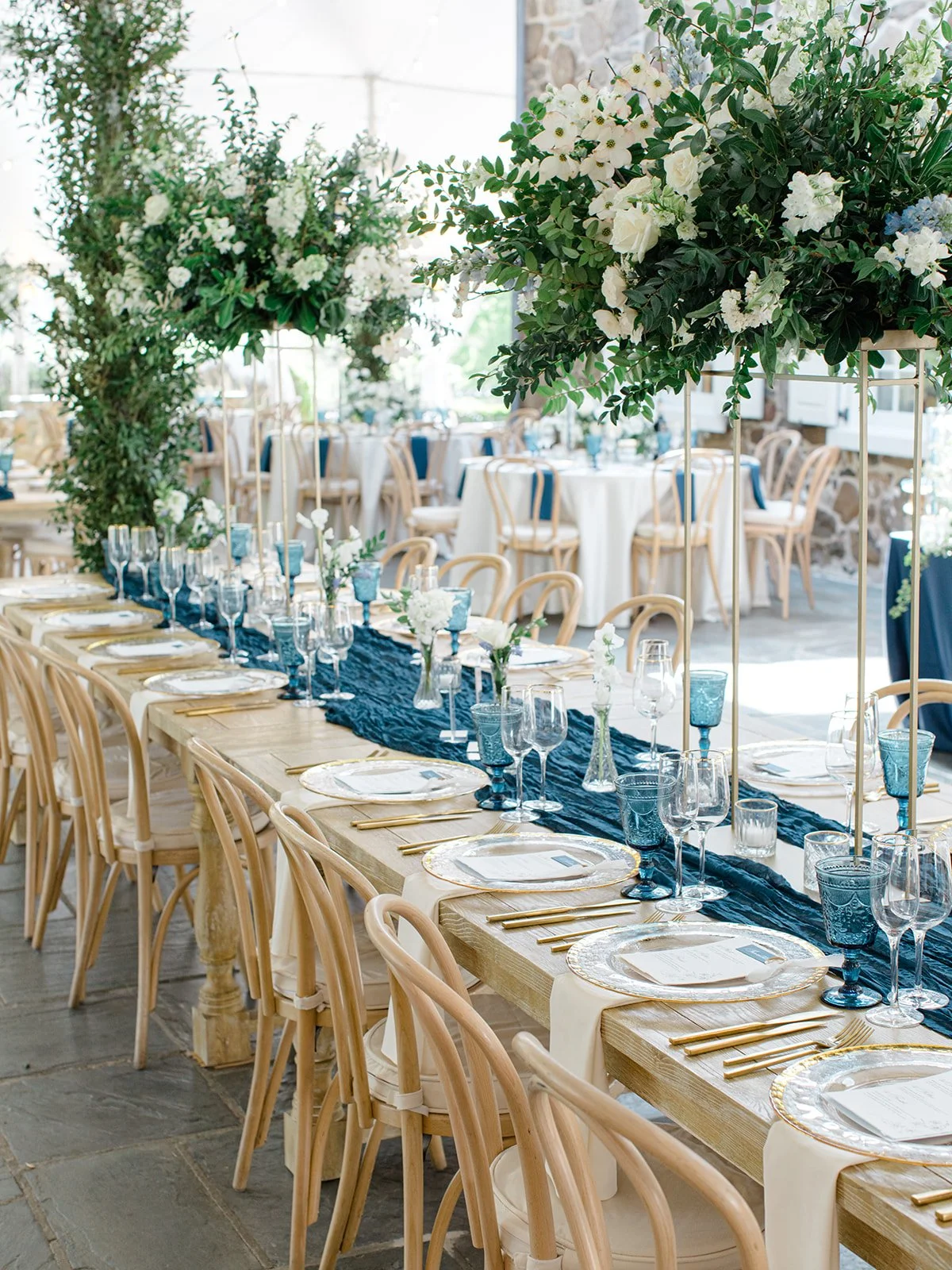 Elegant wedding reception table with white plates, gold flatware, and blue glassware, decorated with tall floral centerpieces of white flowers and green foliage inside a bright space with stone walls.