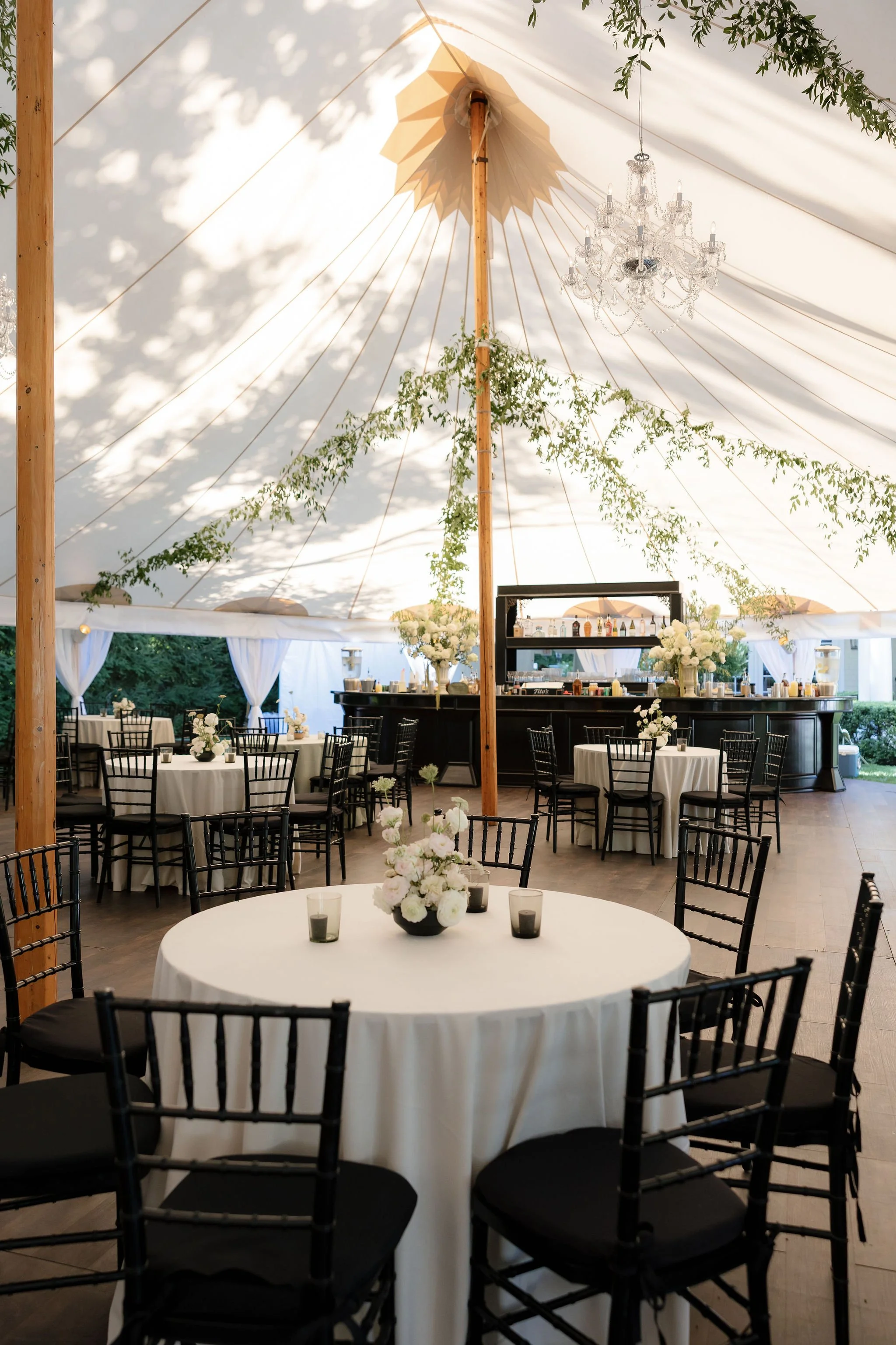 Elegant event tent interior with round tables, black chairs, floral centerpieces, a bar, and a chandelier.