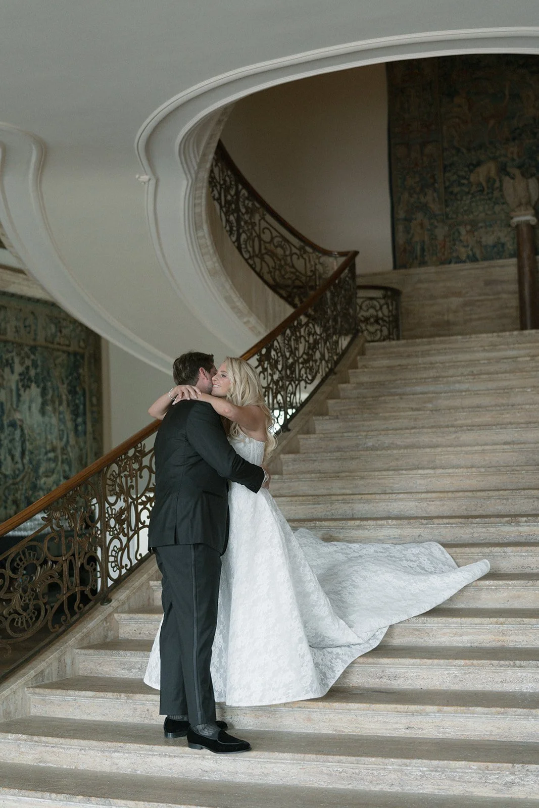 A bride and groom embrace on a grand staircase inside a luxurious venue, with the bride in a white wedding gown and the groom in a black tuxedo.