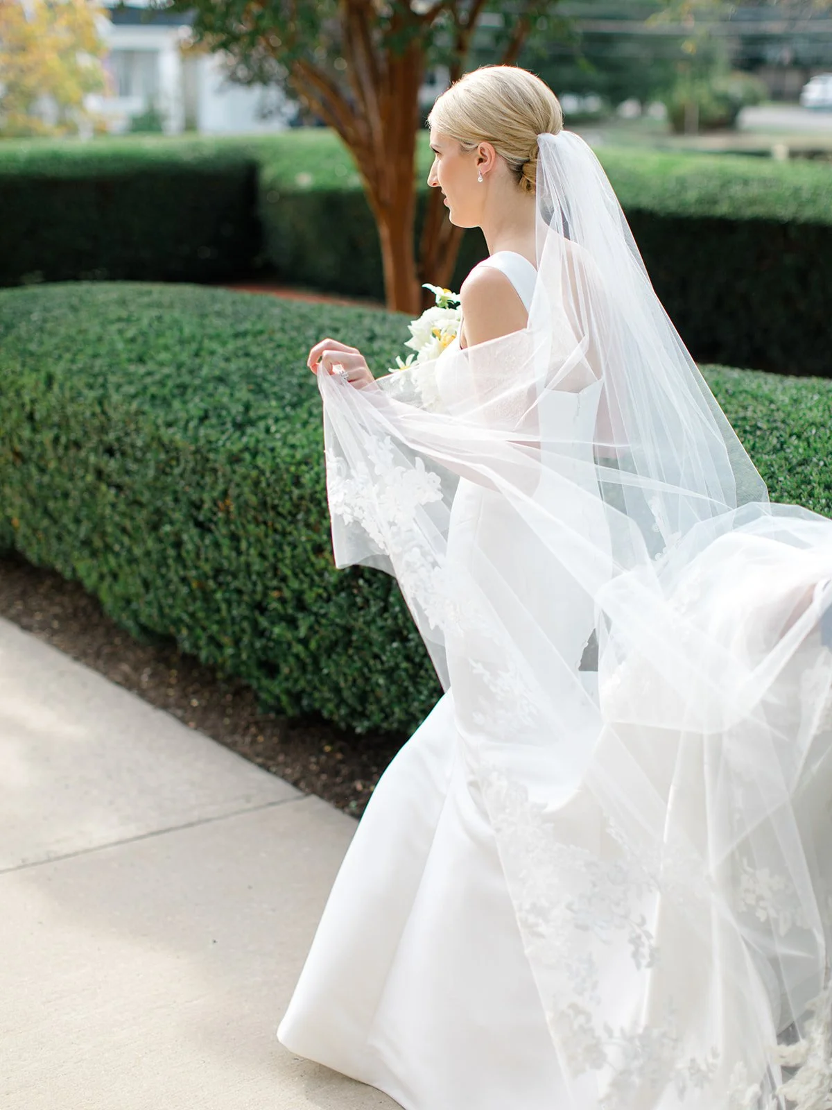 A bride with blonde hair in a bun, wearing a white wedding dress and veil, holding a bouquet, standing near a green hedge.