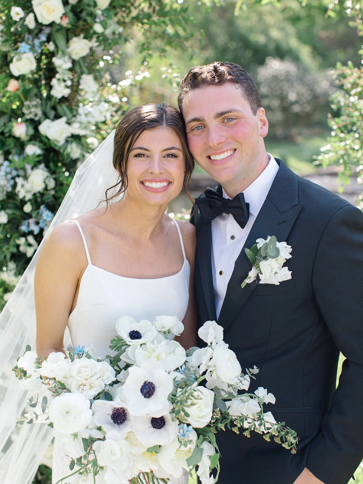 A bride and groom smiling outdoors on their wedding day, surrounded by greenery and flowers, with the bride holding a bouquet of white flowers.