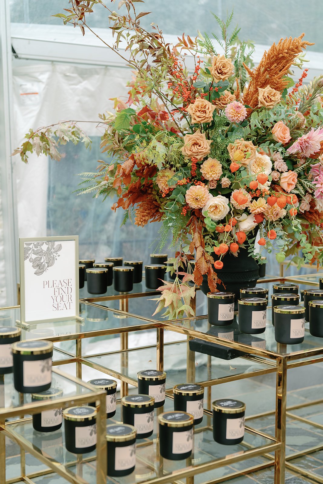 A gold and glass display table with black candles and a large floral arrangement of pink, orange, and peach flowers, greenery, and dried elements, with a sign reading 'Please find your seat' in a white frame.