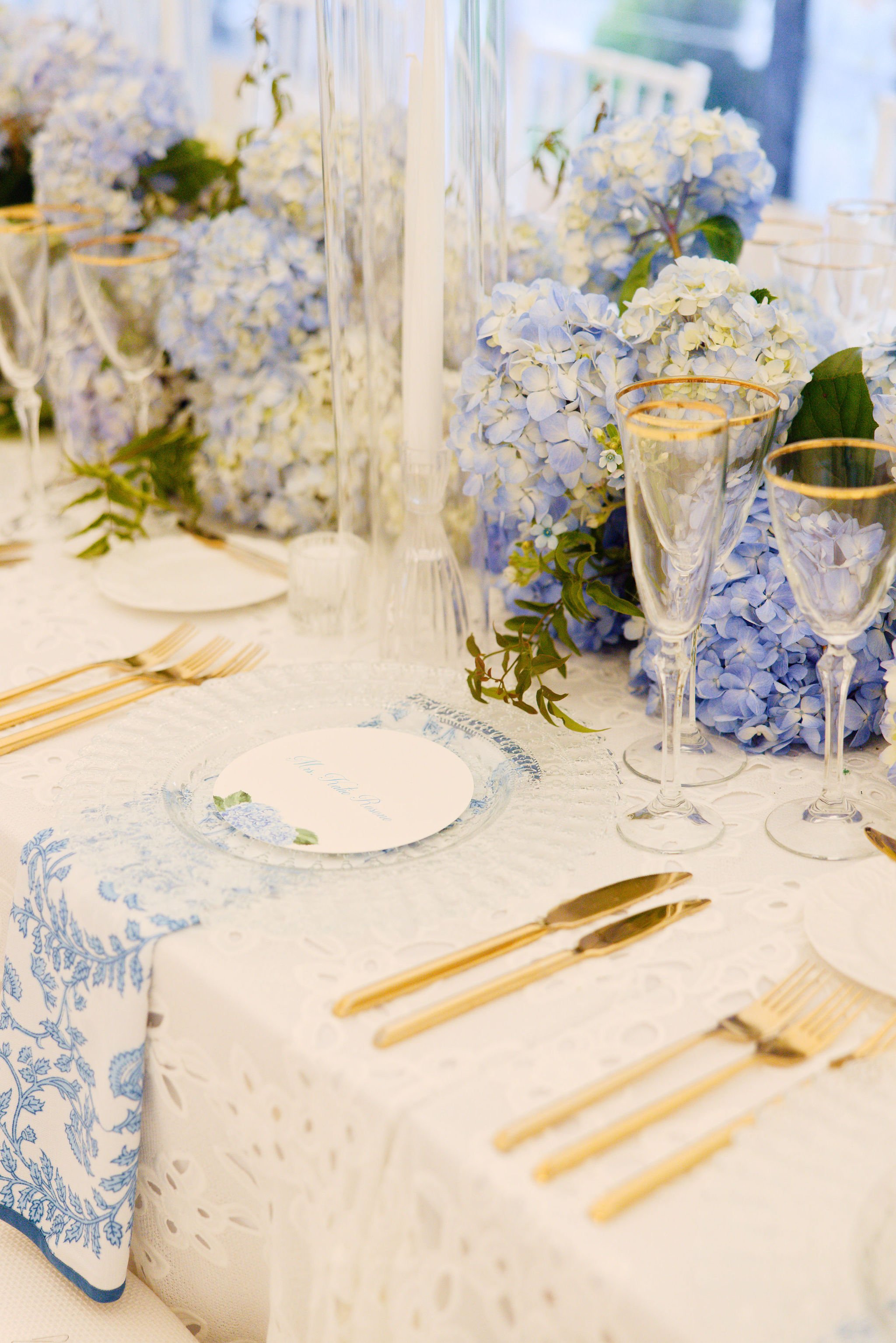 Table setting with gold flatware, clear glassware with gold rims, white lace tablecloth, and blue hydrangea floral centerpiece at a formal event or wedding.