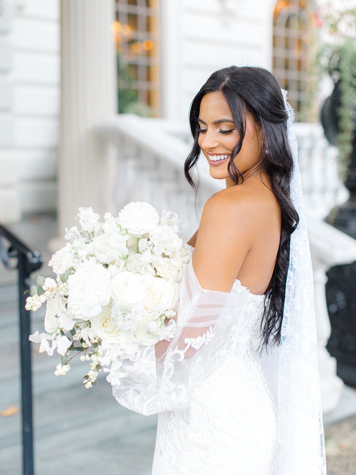 A smiling bride with dark wavy hair wearing a white wedding dress and holding a bouquet of white flowers.