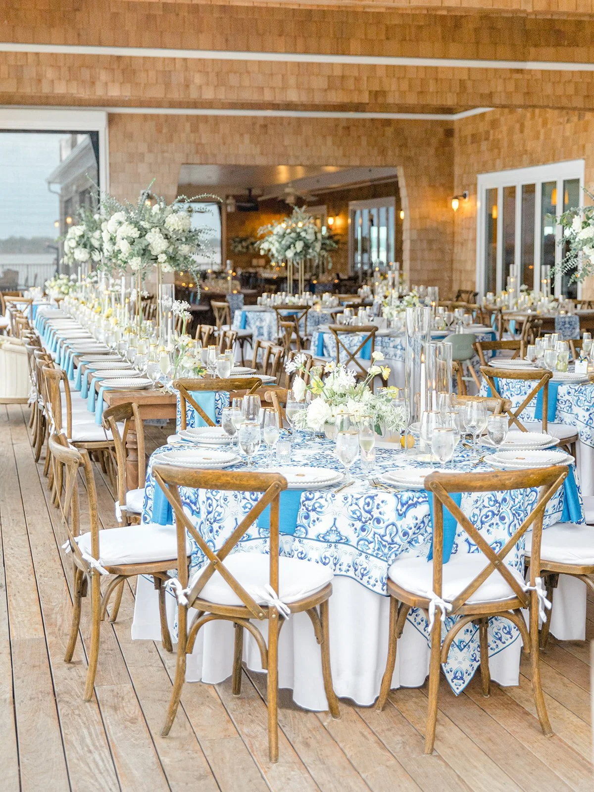 Decorated outdoor dining area for a formal event with multiple round and rectangular tables adorned with white and blue patterned tablecloths, white floral centerpieces, glassware, and candles. Wooden chairs with white cushions surround the tables, a