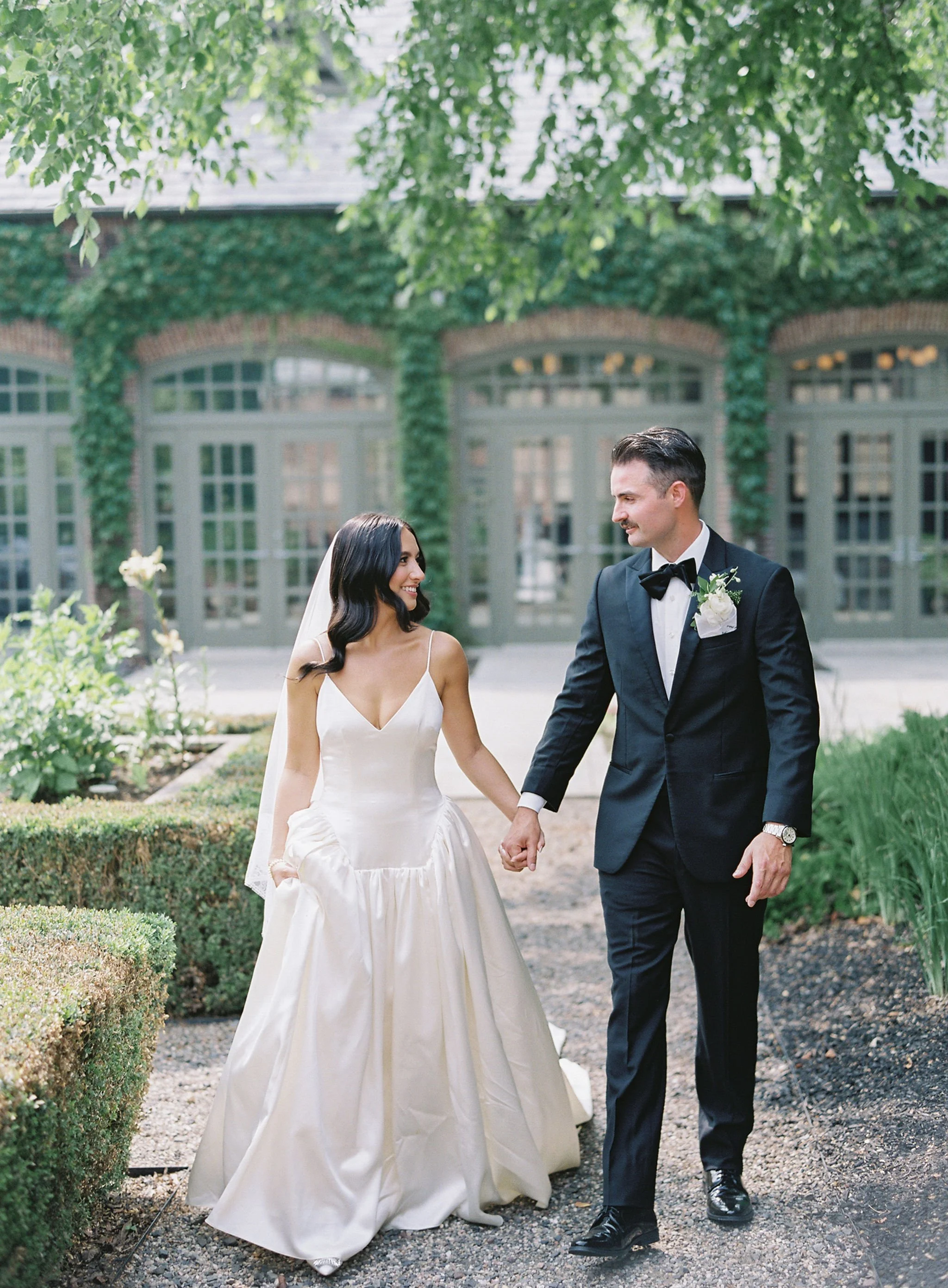 A bride and groom walking hand in hand outdoors, holding hands and smiling at each other, with greenery and a building with large windows in the background.