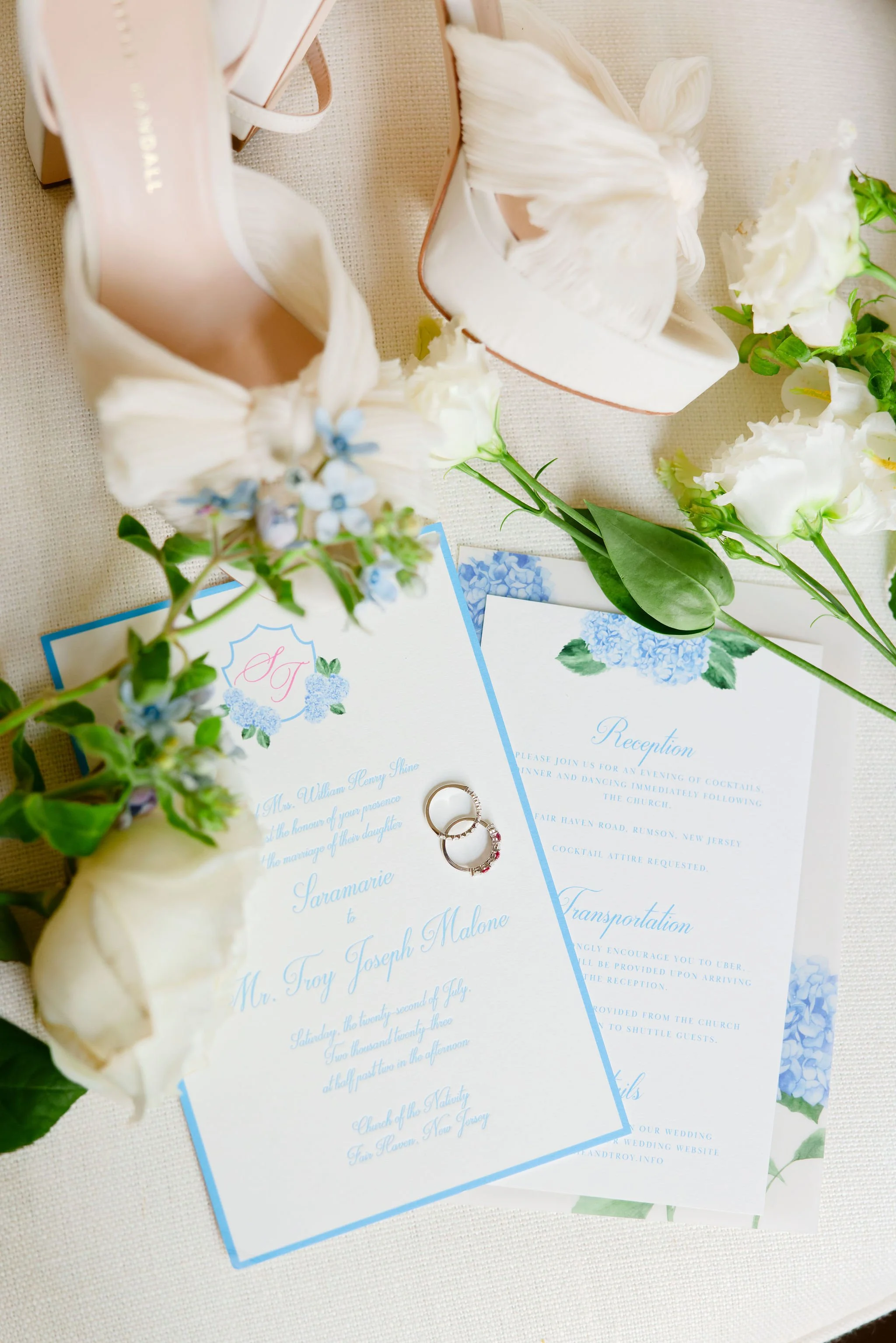 Wedding invitation with rings, flowers, and white heels placed on a table.