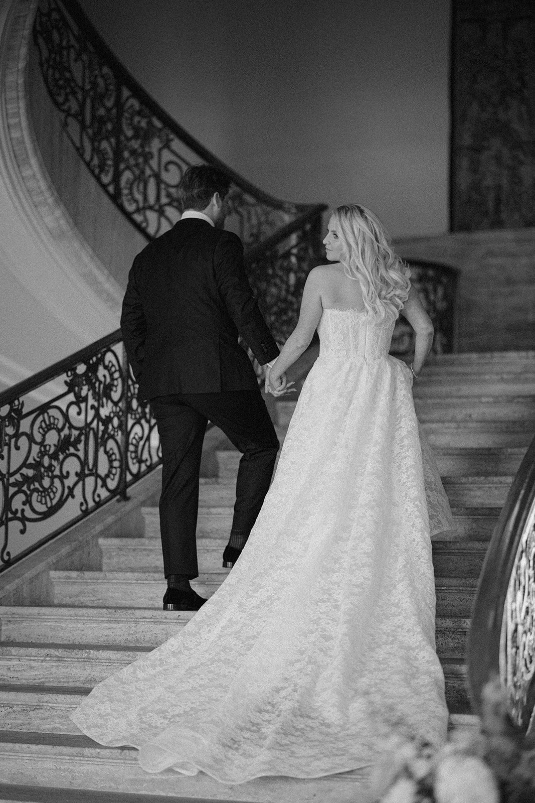 A black-and-white photo of a bride and groom holding hands and ascending a staircase indoors. The bride is wearing a long, strapless wedding gown, and the groom is dressed in a black suit. The staircase has ornate wrought iron railing and a patterned