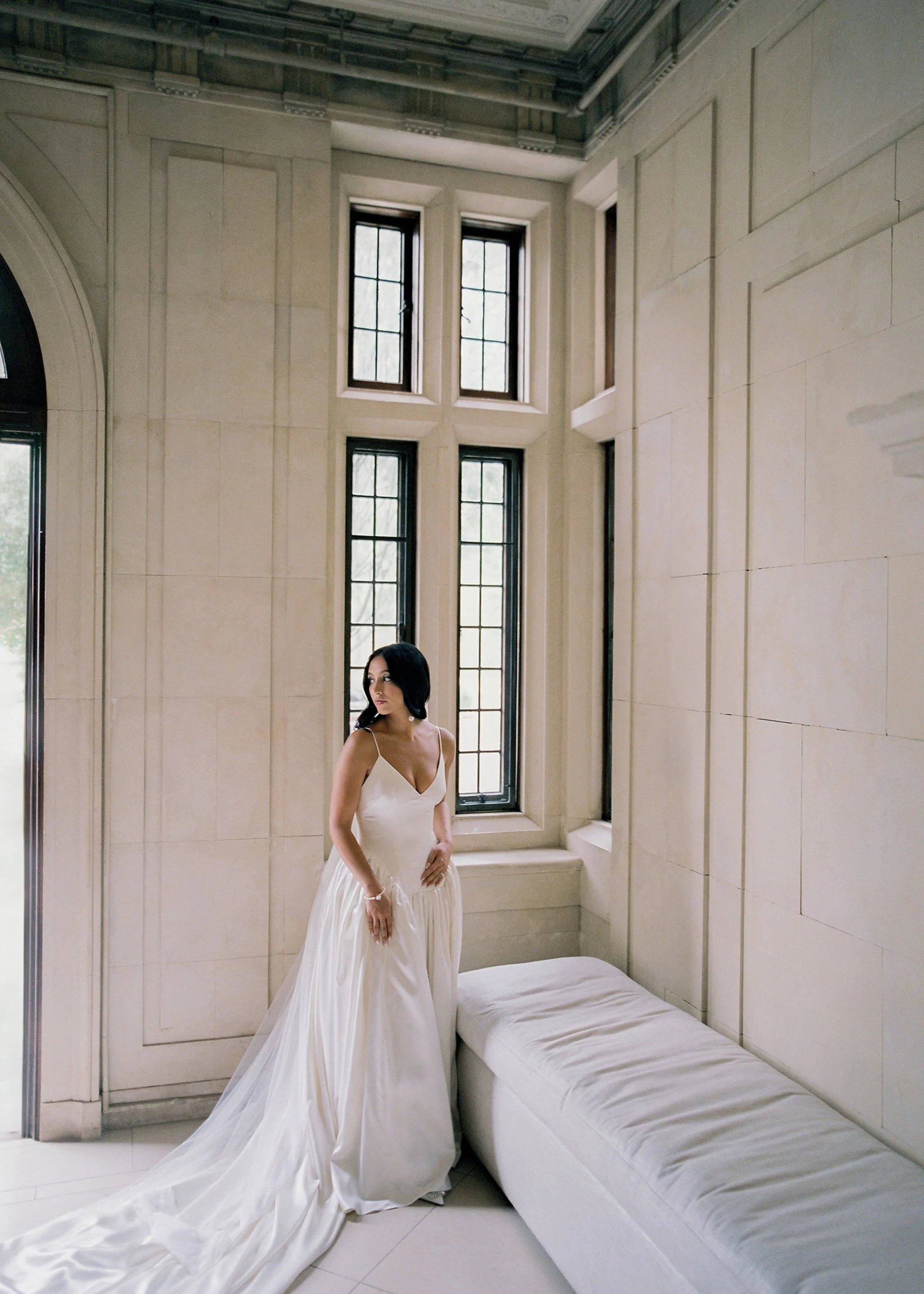 A woman in a white wedding dress standing near a window in a bright, elegant room with tall, divided windows and light-colored walls.