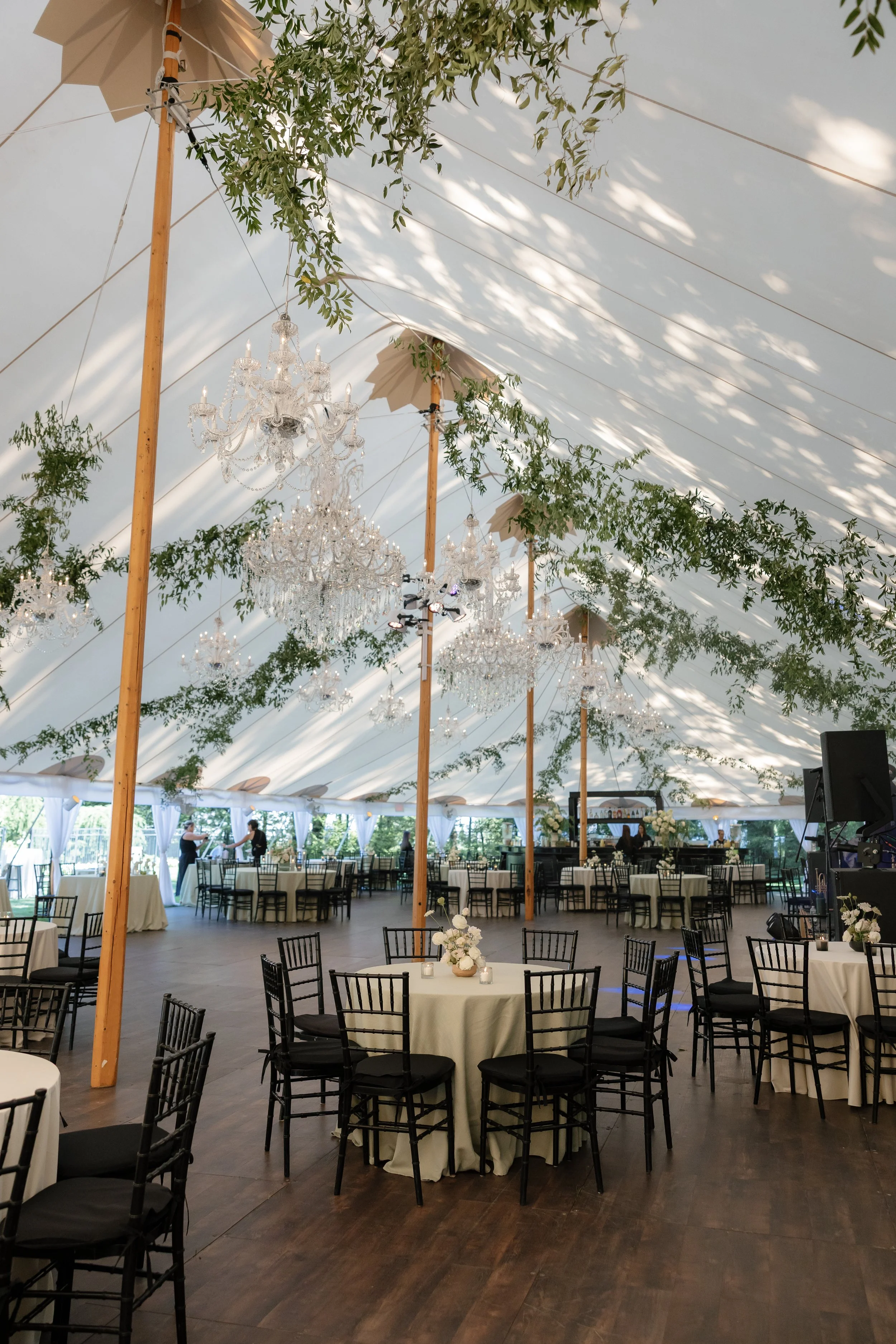 Elegant event tent decorated with chandeliers, greenery, and black chairs around tables with white tablecloths for a formal occasion.
