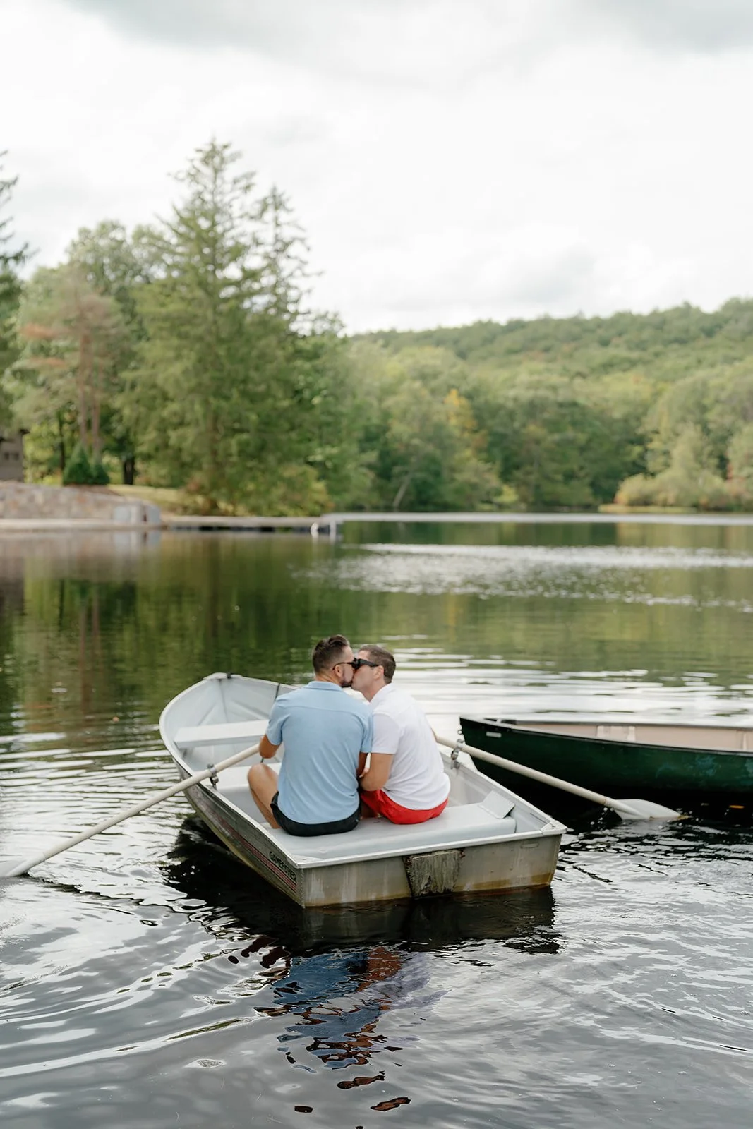 Two men sitting in a small boat on a lake, kissing, during daytime with trees and hills in the background.