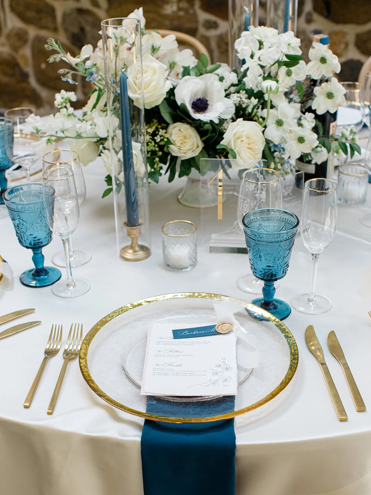 Elegant table setting with a floral centerpiece, blue glasses, gold flatware, a clear glass plate with a menu, and navy blue napkin at a formal event.
