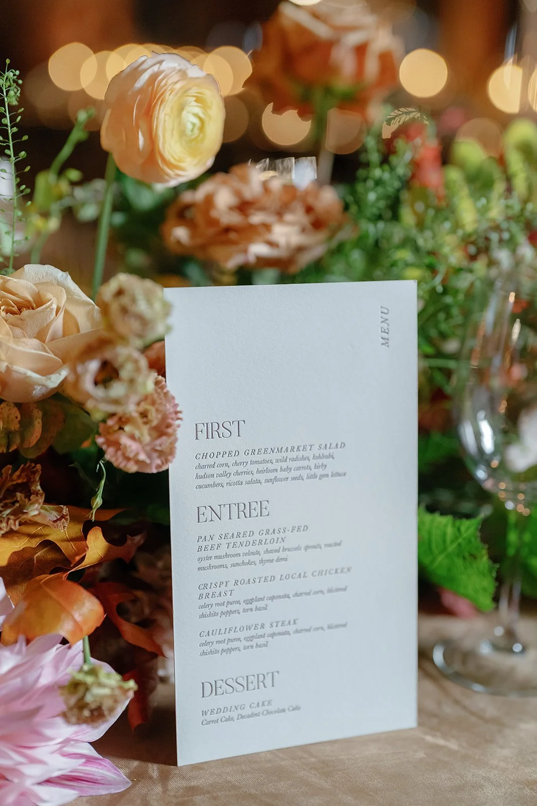 A white menu card placed among a variety of peach, pink, and orange flowers on a wooden table.