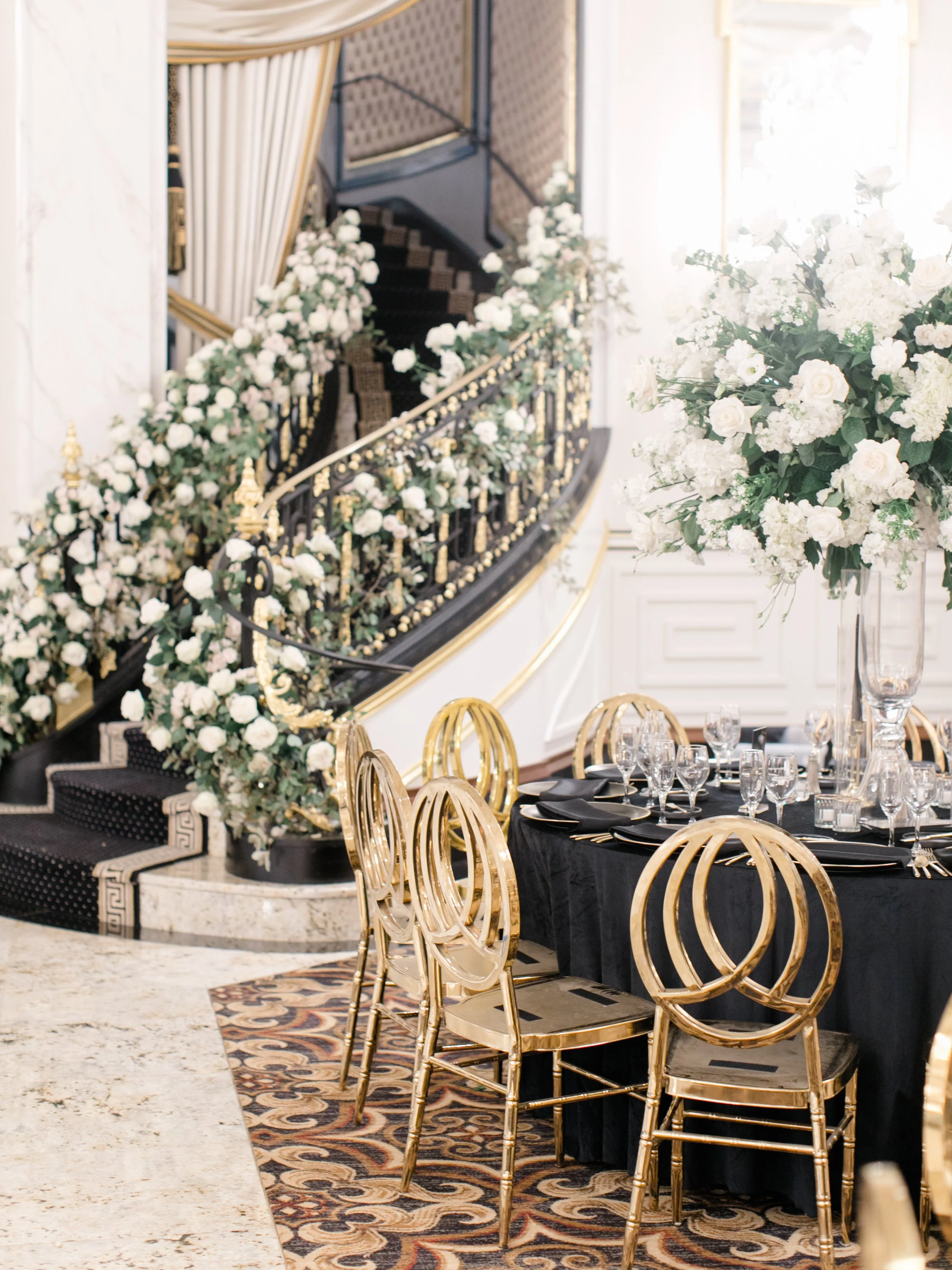 Elegant banquet table with black tablecloth, gold chairs, and tall floral centerpiece of white roses and greenery, set in a luxurious venue with a curved staircase decorated with white flowers and gold accents.