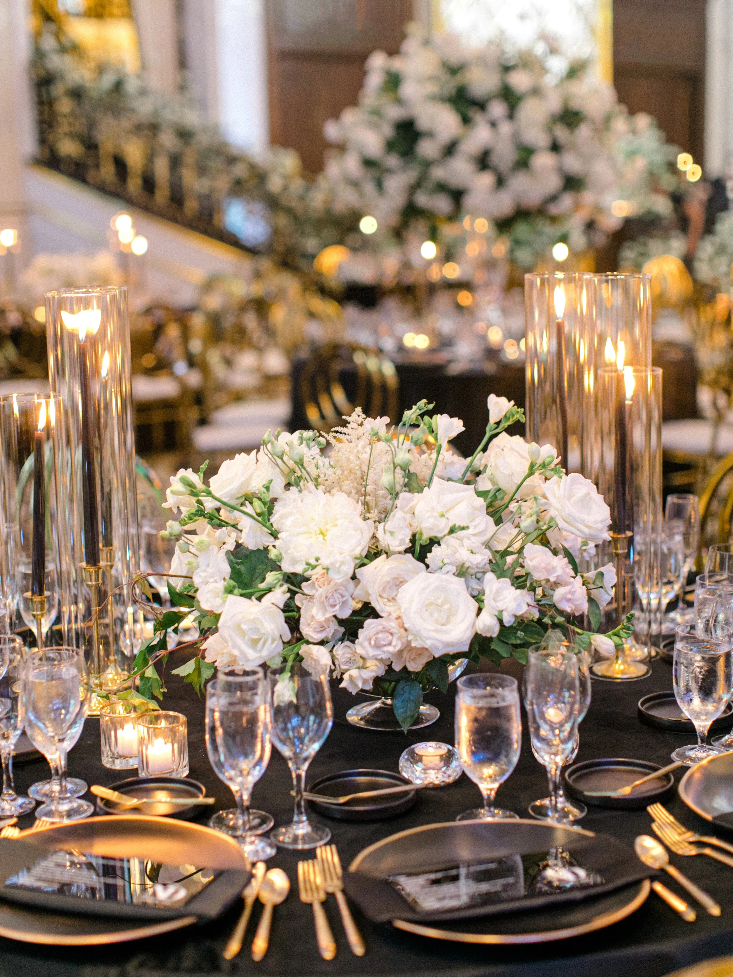 Elegant banquet table with white floral centerpiece, tall glass candle holders, and gold utensils in a decorated event hall.