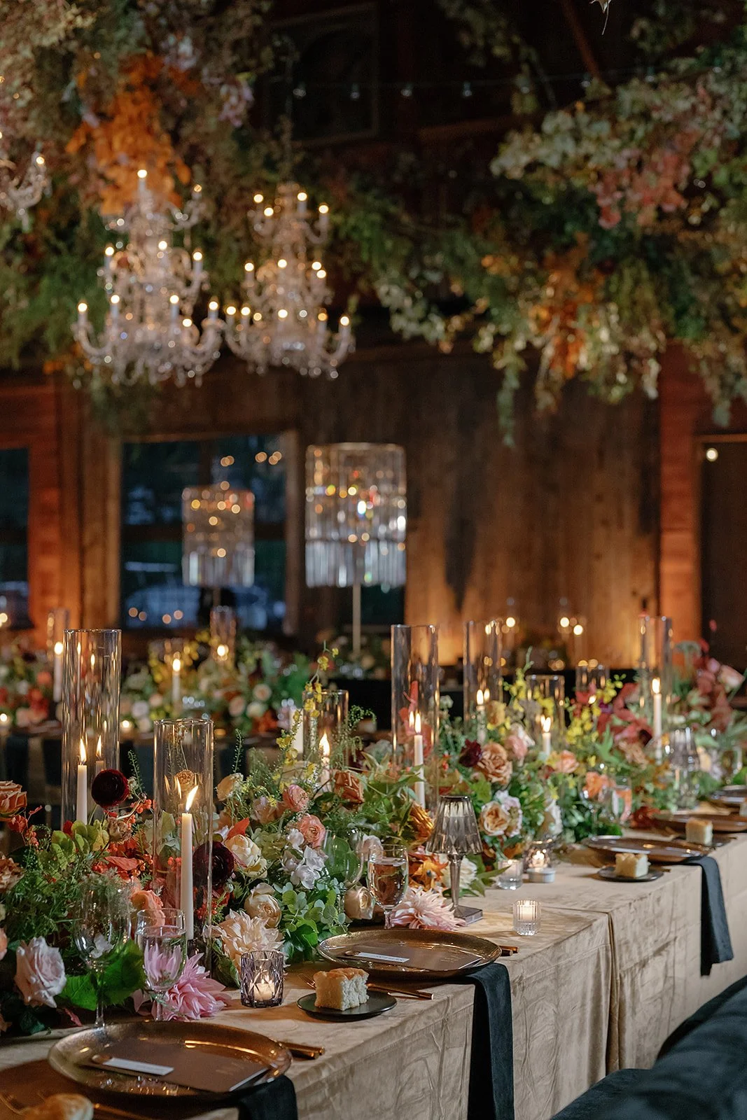 Elegant wedding reception table with floral centerpieces, candles, and crystal chandeliers in a rustic wooden venue.
