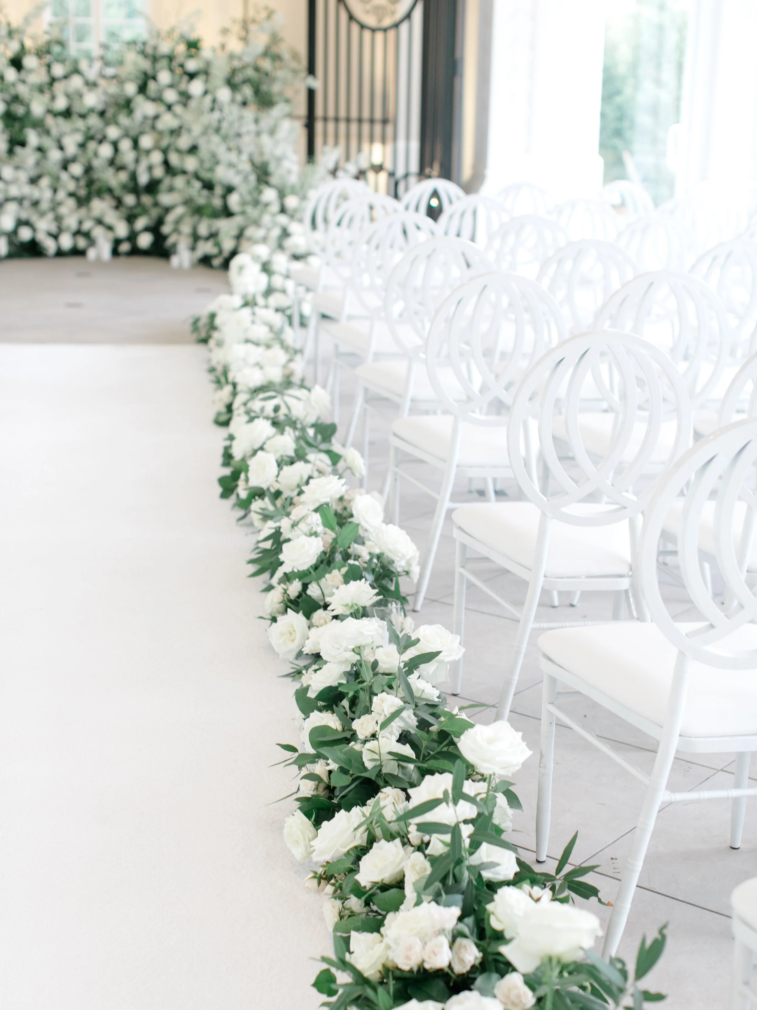 White wedding chairs lined up with a white floral aisle runner and white floral arrangements on either side of the aisle.