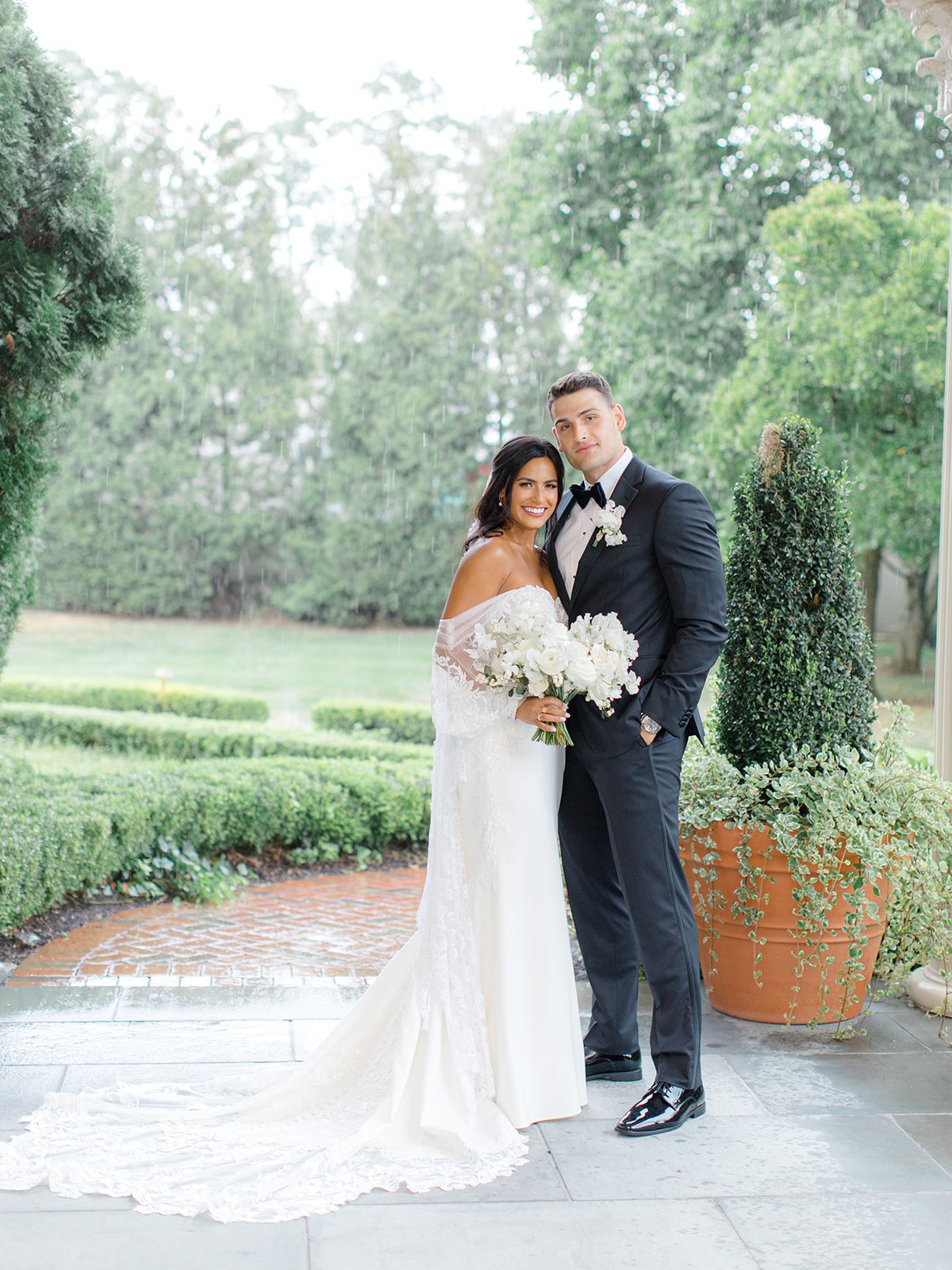 A bride and groom stand together outdoors on a rainy day. The bride is in a white off-shoulder wedding gown holding a bouquet of white flowers. The groom is in a black tuxedo with a bow tie and boutonniere. They are smiling and embracing in front of 