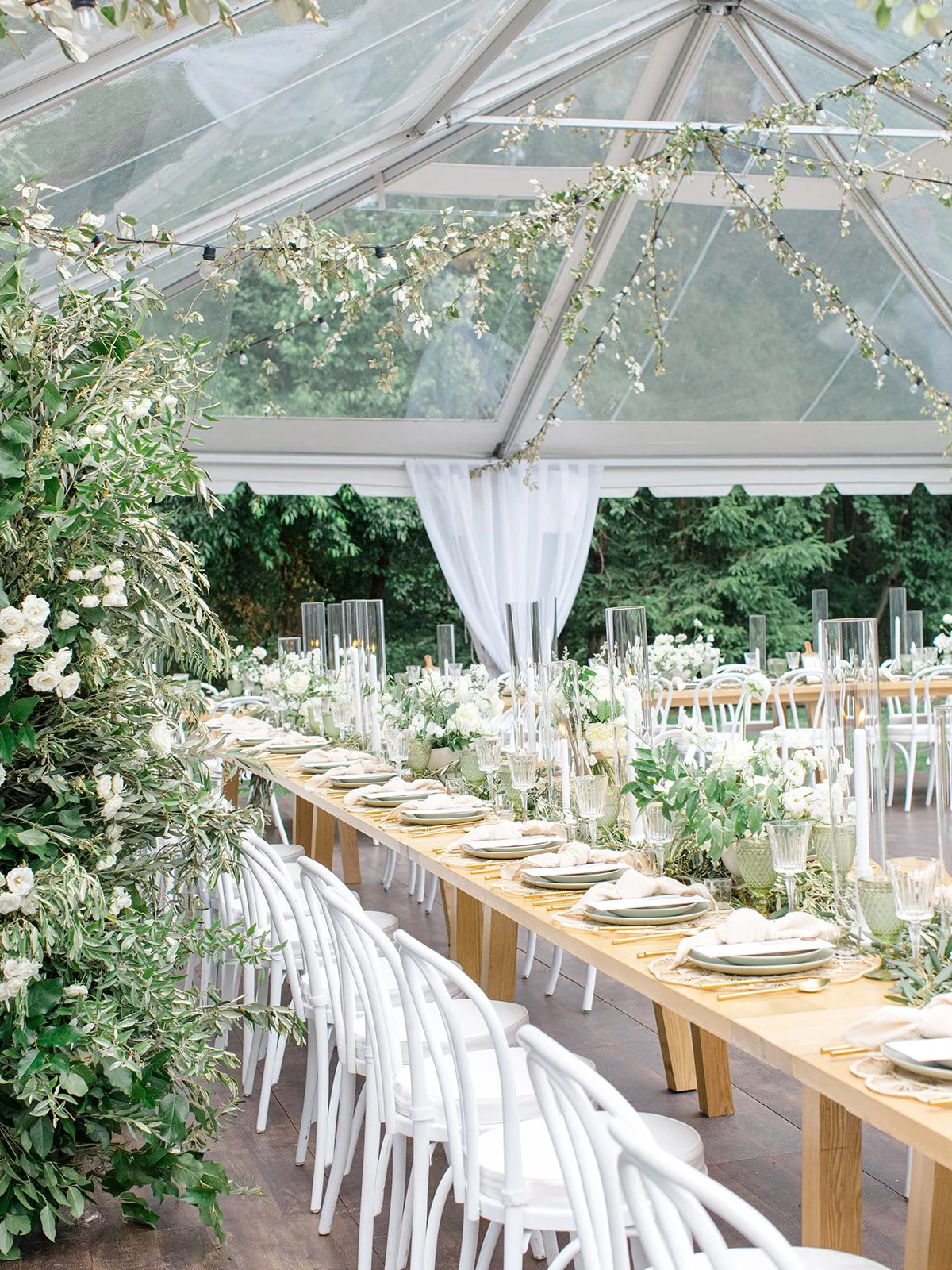 Elegant outdoor dining setup under a glass canopy with white flowers and greenery, featuring a long wooden table with white chairs, glassware, plates, and decorative candles for a special event or wedding reception.