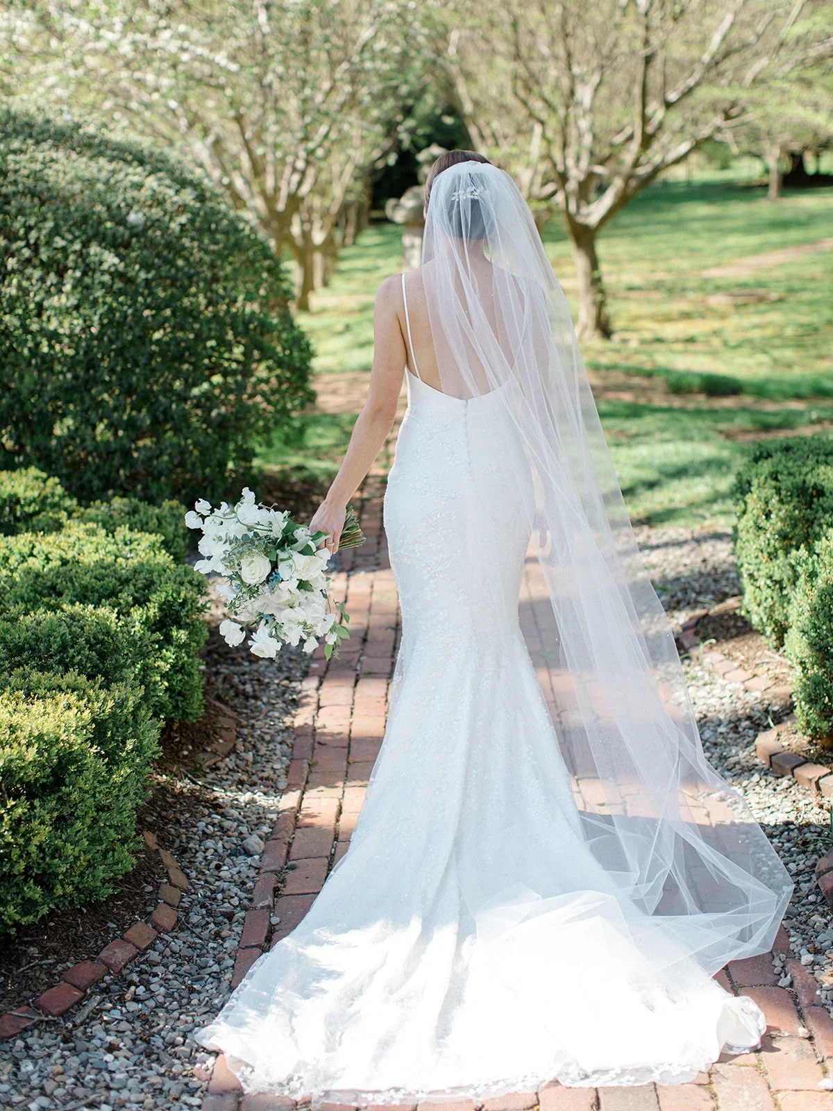 Bride in a white gown and veil walking on a brick pathway in a garden, holding a bouquet of white flowers.