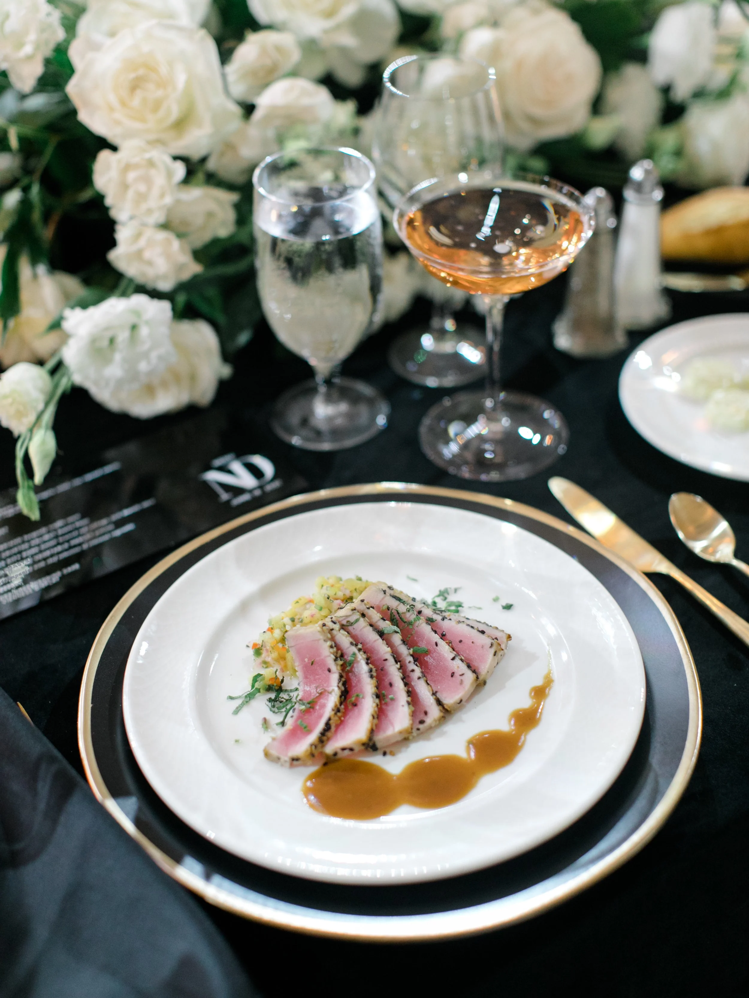A fancy dinner plate with seared tuna steak garnished with herbs, served with a sauce and mashed potatoes, set on a black and gold-rimmed plate on a black tablecloth with silverware and glassware, and a floral centerpiece of white roses in the backgr