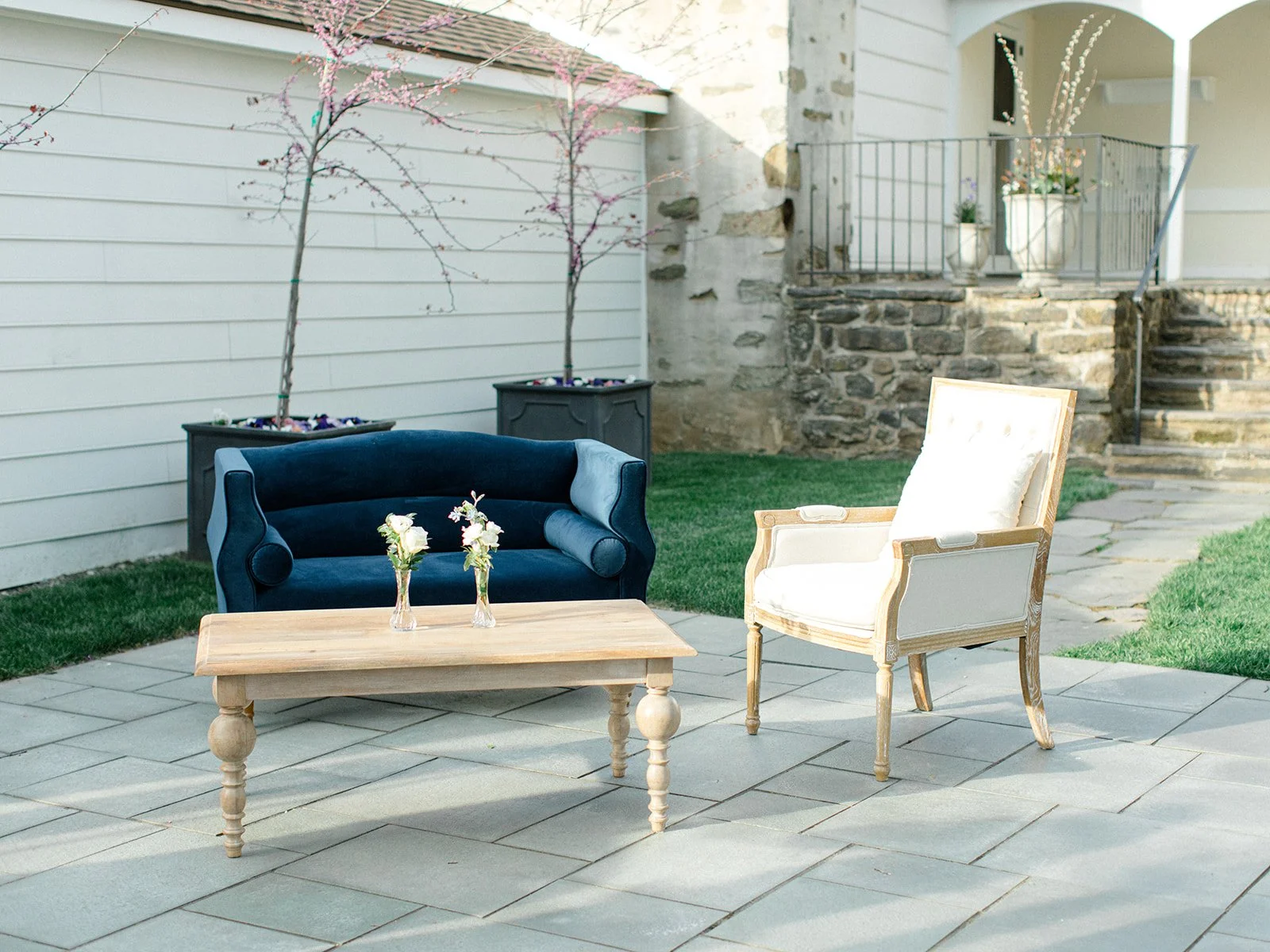 Outdoor patio with a blue velvet sofa, a cream-colored armchair, a wooden coffee table with two vases of flowers, two pink flowering trees in pots, stone stairs leading to an upper level, and a house with white siding and a stone wall.