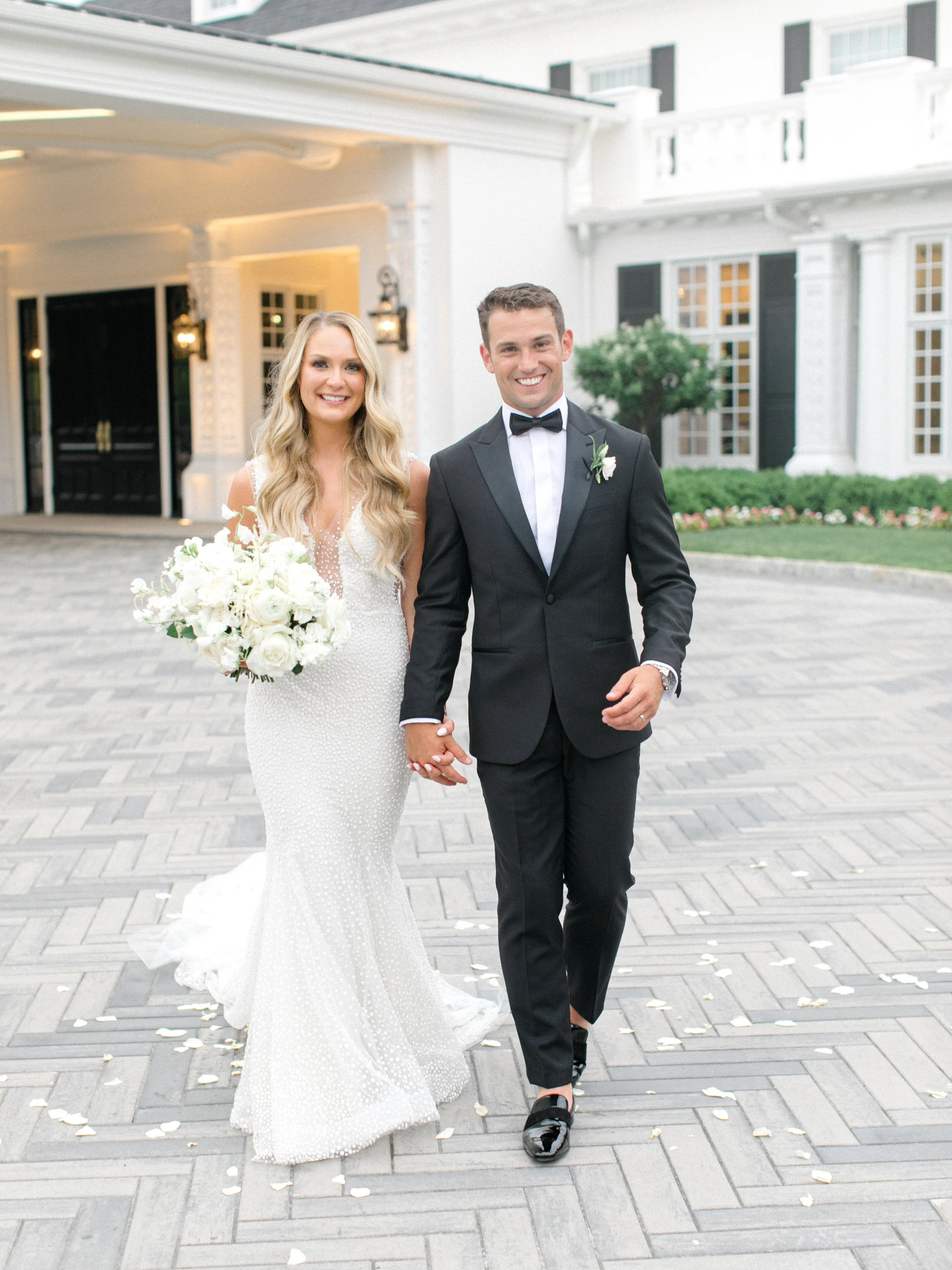 A newlywed couple holding hands and walking outside a white building with black shutters. The bride is in a white wedding dress holding a bouquet of white flowers, and the groom is in a black tuxedo with a bow tie.