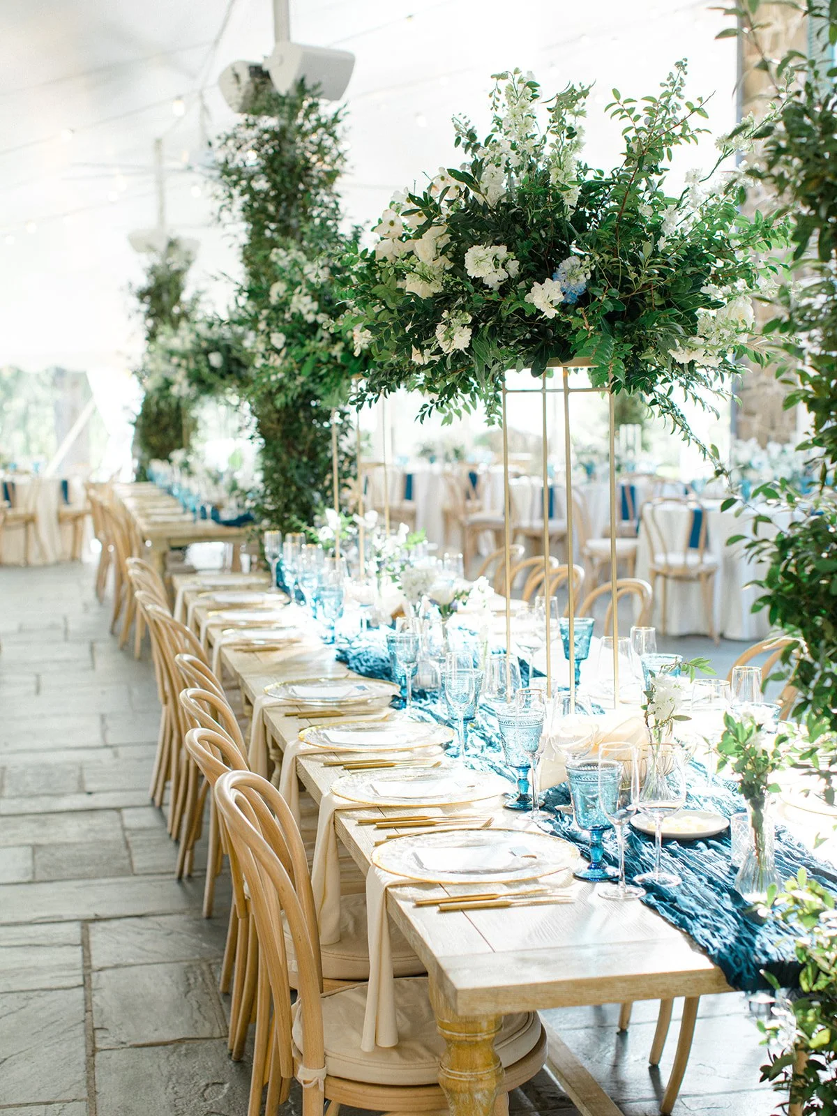 Elegant banquet table set with white plates, gold flatware, clear and blue glassware, white floral arrangements, and lush greenery in an airy tent with natural light.