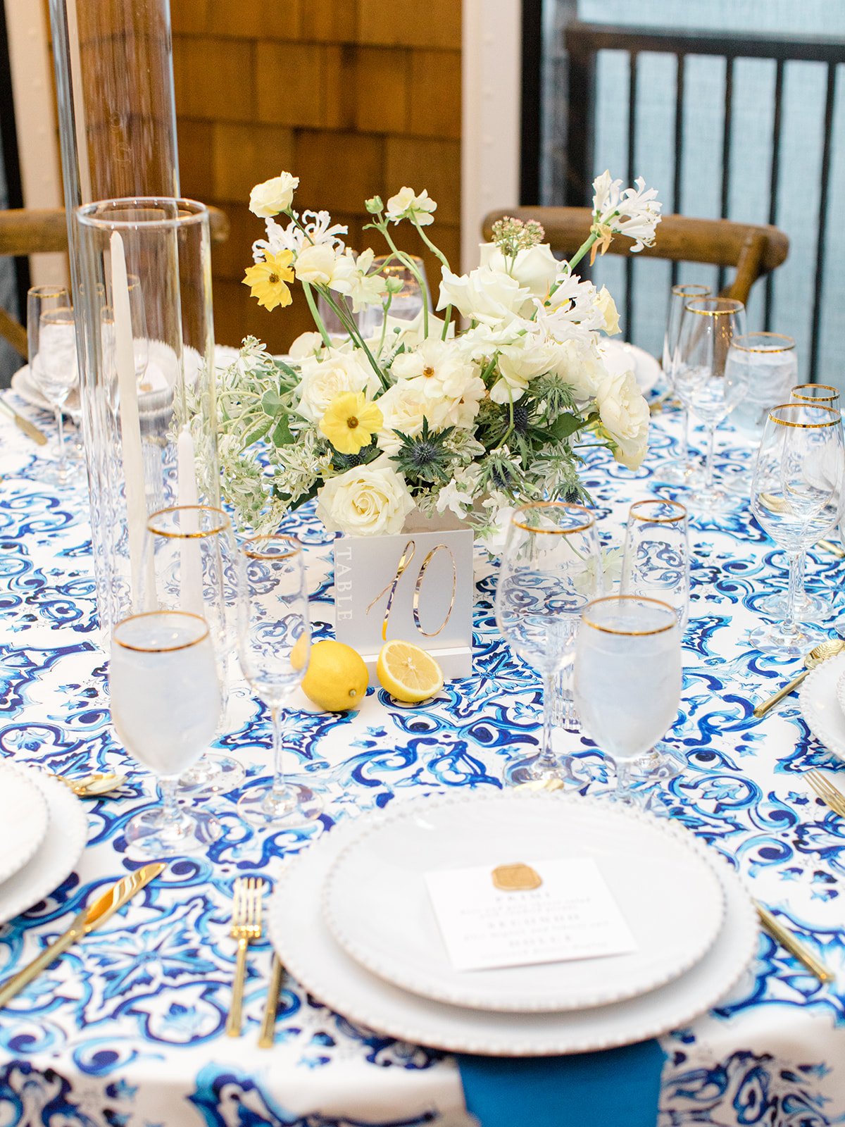 Table decorated with a blue and white patterned tablecloth, a floral centerpiece with white and yellow flowers, and elegant glassware for a formal event.