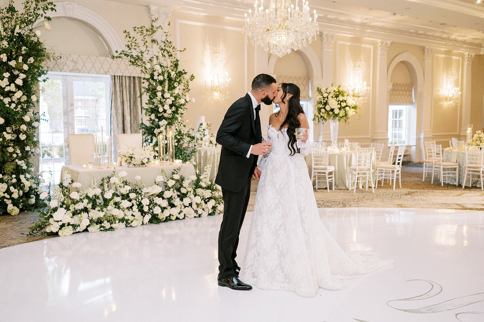 A bride and groom share a kiss at their wedding reception in an elegant, decorated ballroom with white floral arrangements and large windows.