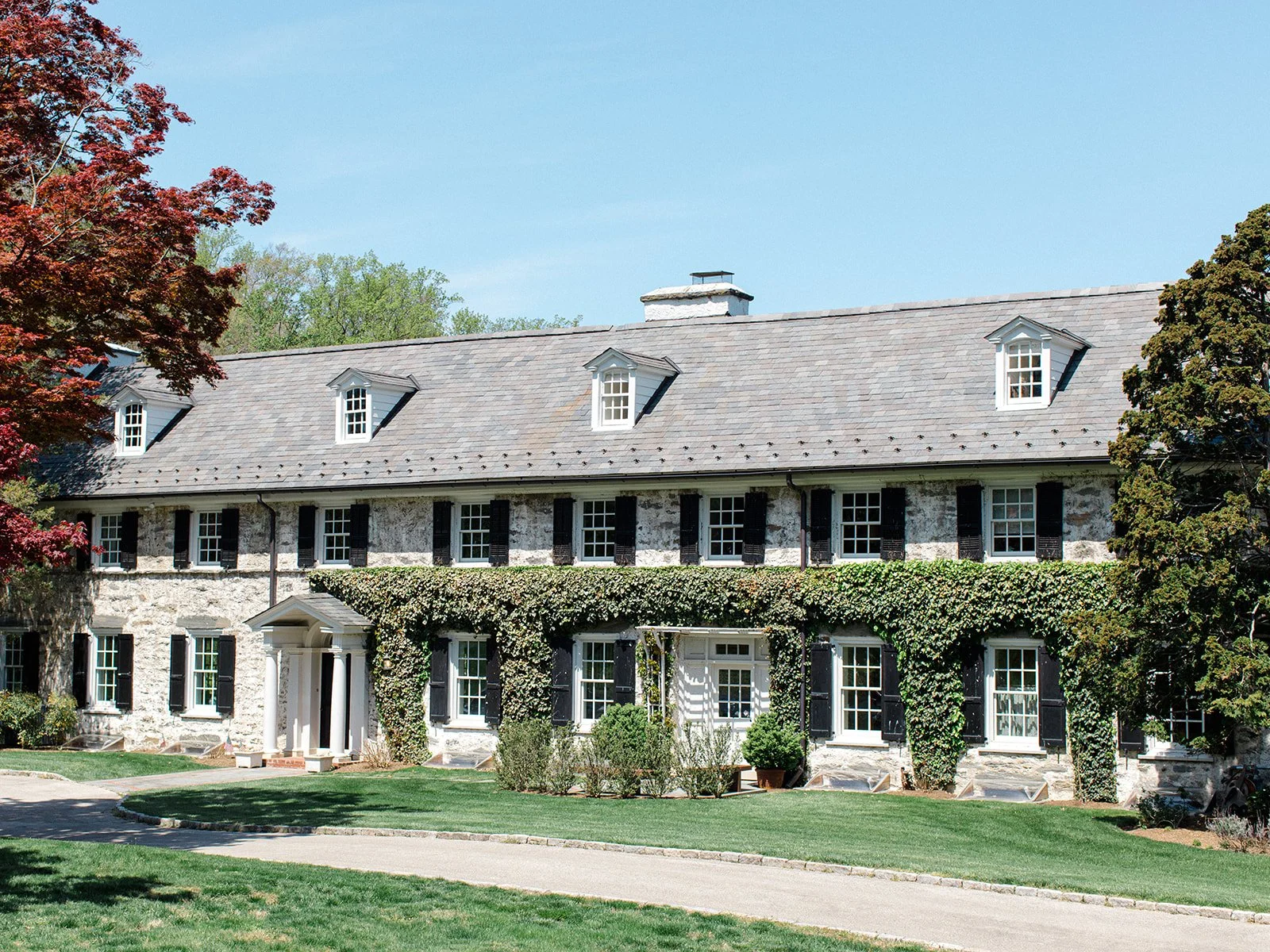 A large, three-story stone house with a gray shingle roof, black shutters, and white-framed windows, partially covered with green ivy. The house has a front porch with white columns and a well-maintained lawn with trees on either side.