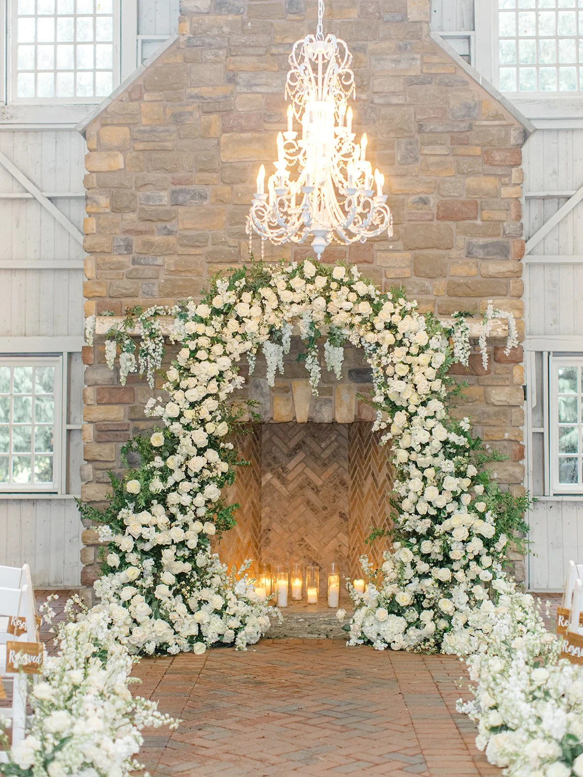 A wedding ceremony setup inside a rustic building with a stone fireplace, decorated with a large floral arch of white roses and greenery, in front of the fireplace are candles on the floor. A chandelier hangs from the ceiling above.