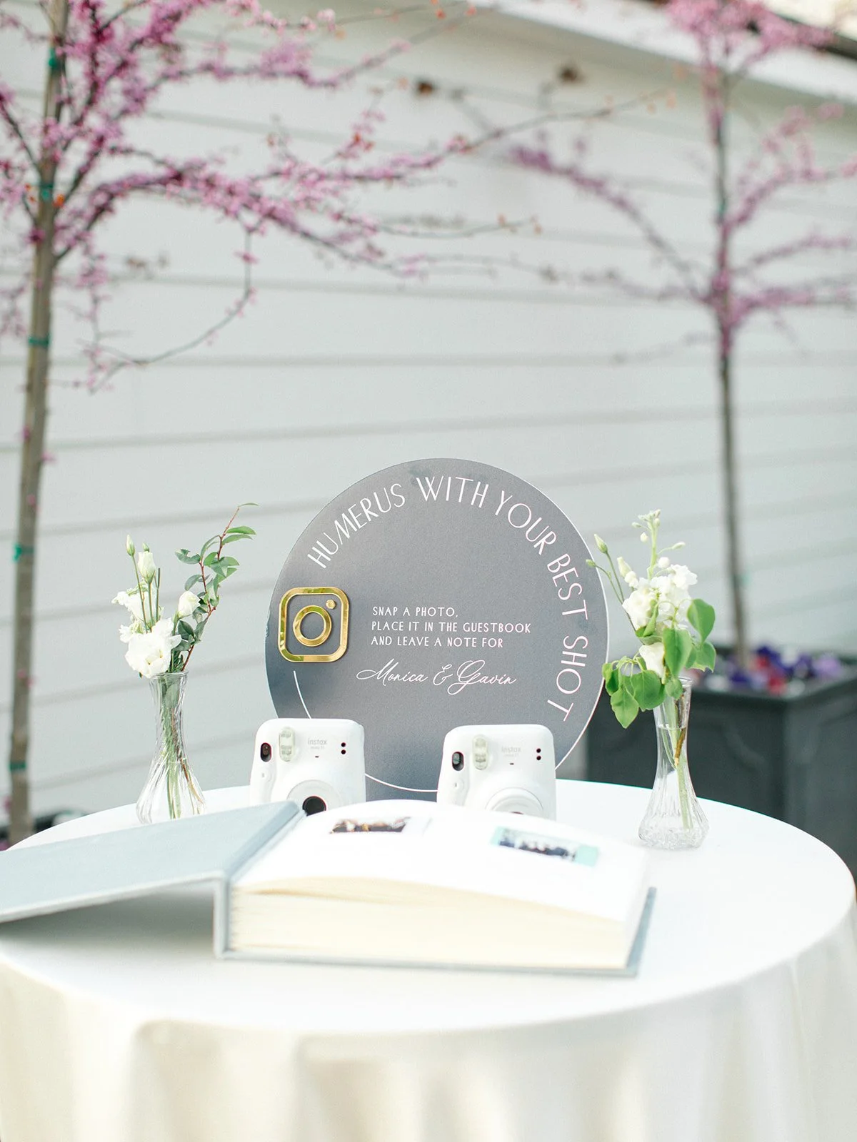 Table with cameras, photo book, flower vases, and a guestbook in front of a pink blossoming tree.