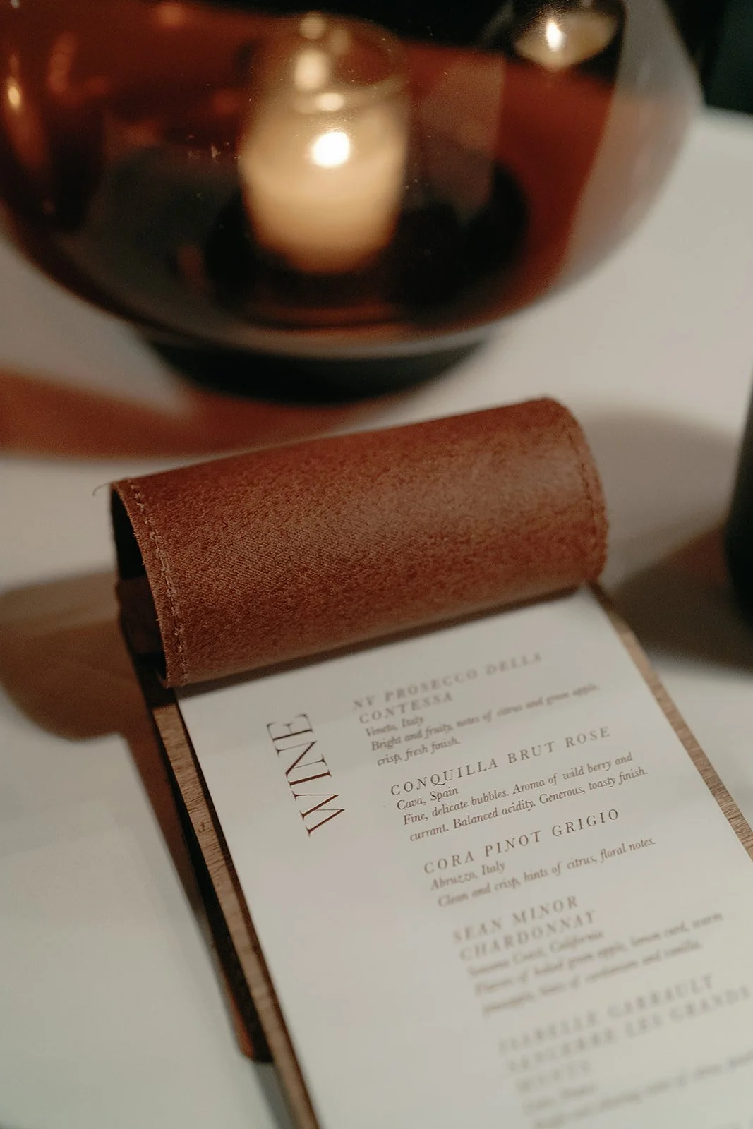 A wine menu with a brown leather cover and a list of wines, positioned on a white table, with a large dark glass candle holder in the background.